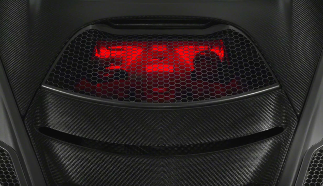 Close-up detail shot of the red, glowing light and honeycomb grille on the rear deck of a McLaren supercar, framed by carbon fiber bodywork.