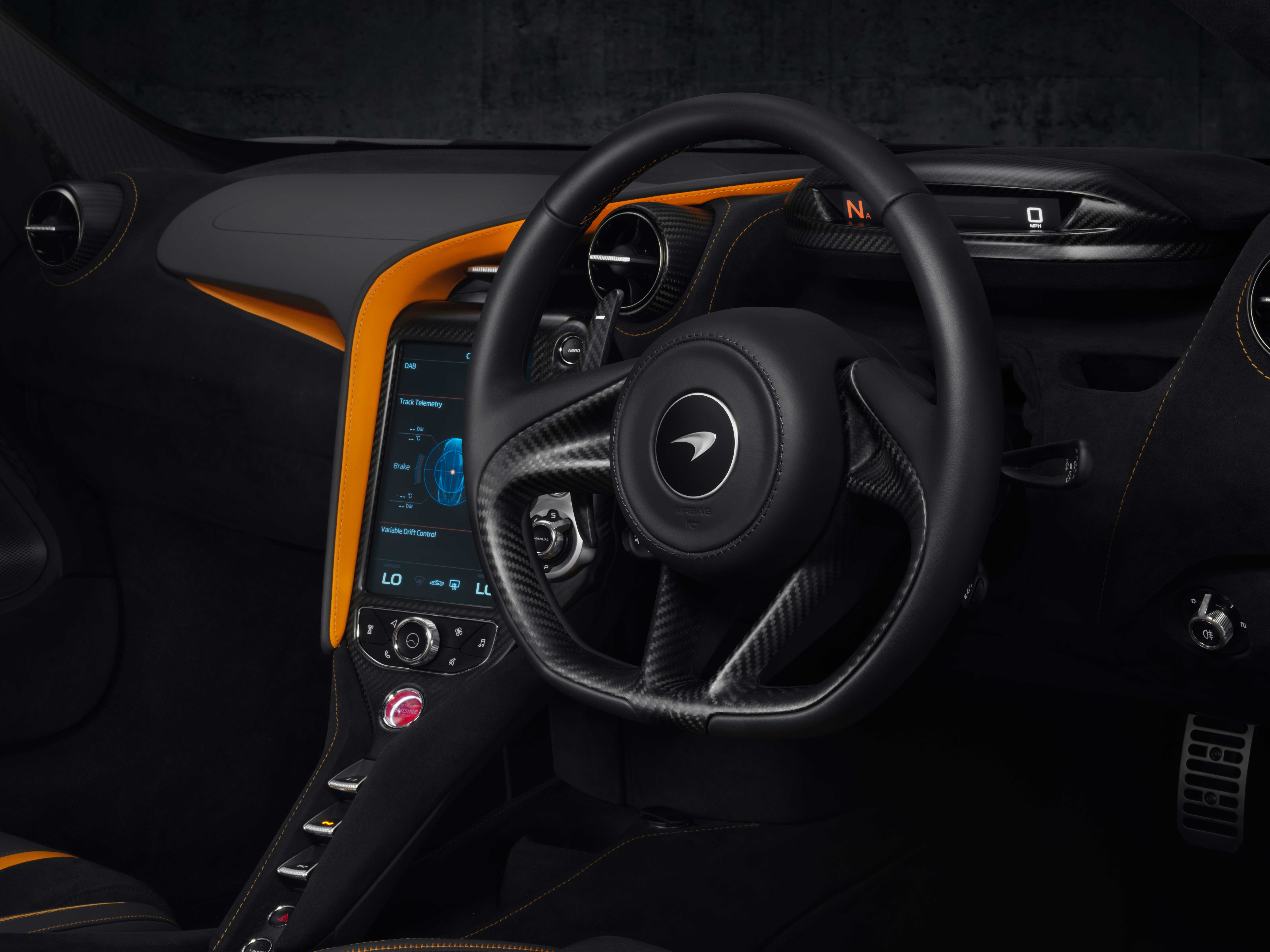 A close-up of the interior of a McLaren 720S, showing the carbon fiber steering wheel with the brand logo, the central infotainment screen, and the orange and black dashboard trim.