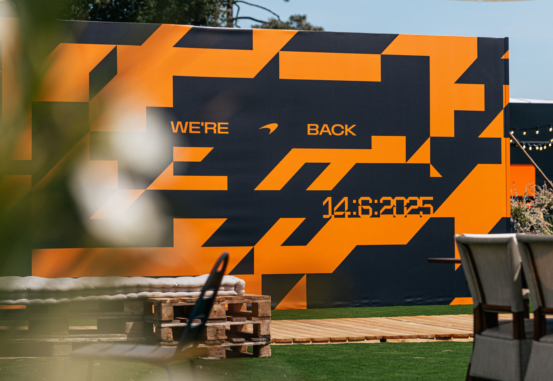 A large black and orange geometric patterned wall with the text "WE'RE BACK" and the date "14:6:2025," used for a McLaren brand event.