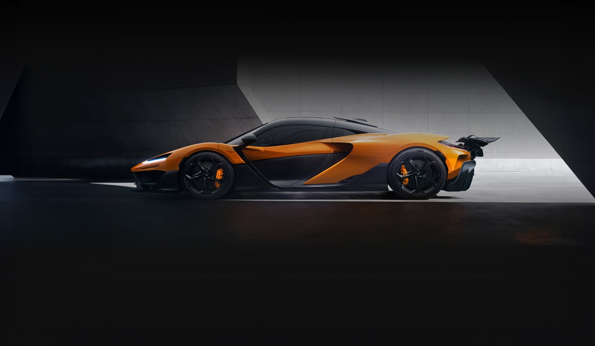 A stunning orange and black McLaren W1 is shown from a side profile in a dark, moody studio. The image highlights the car's sleek lines, bright brake calipers, and lightweight wheels.