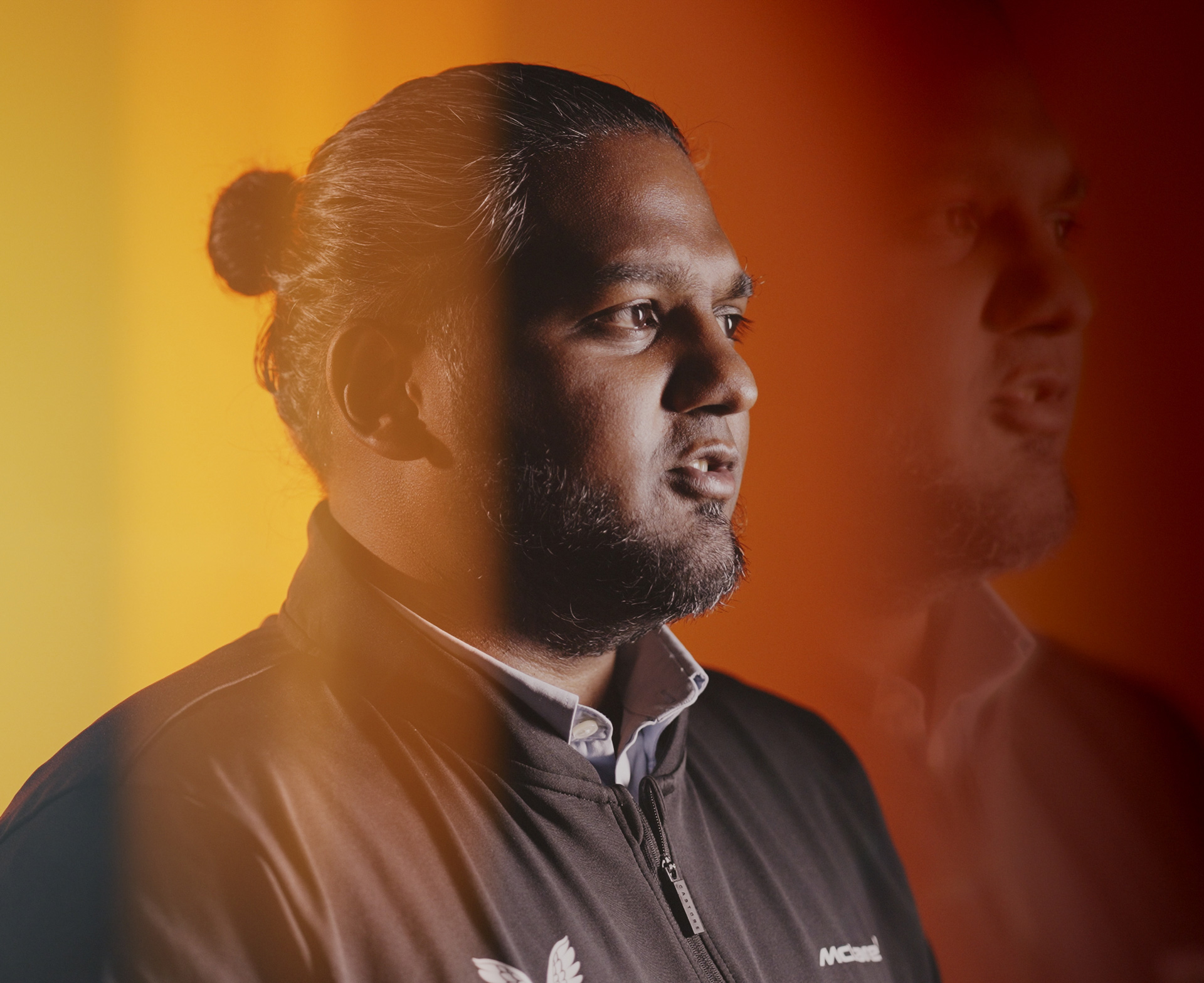 professional headshot of a man with a beard and hair in a bun, looking thoughtfully to the side against a blurred, orange and yellow background.