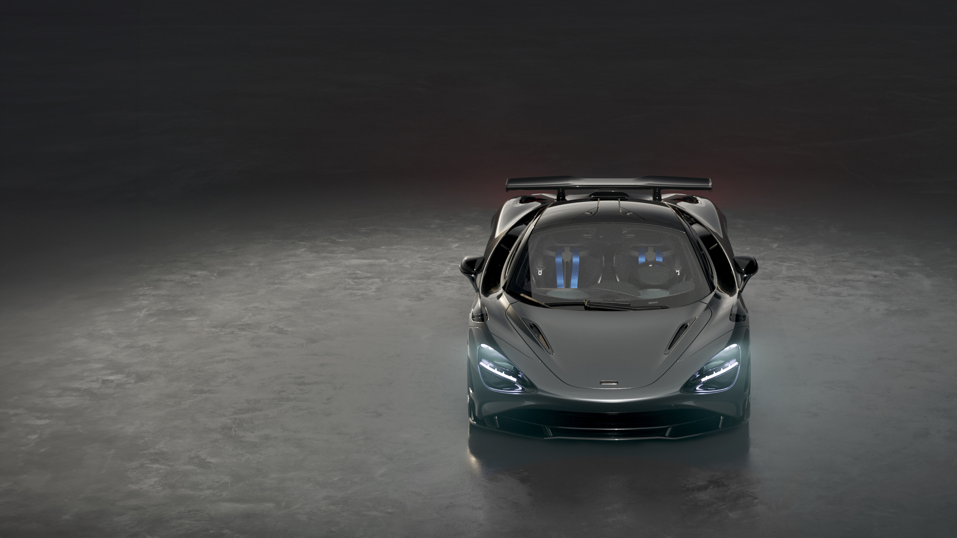 A dark grey McLaren 750S Le Mans special edition supercar is shown from a front-on view in a dark studio, with its headlights on and a prominent rear spoiler.