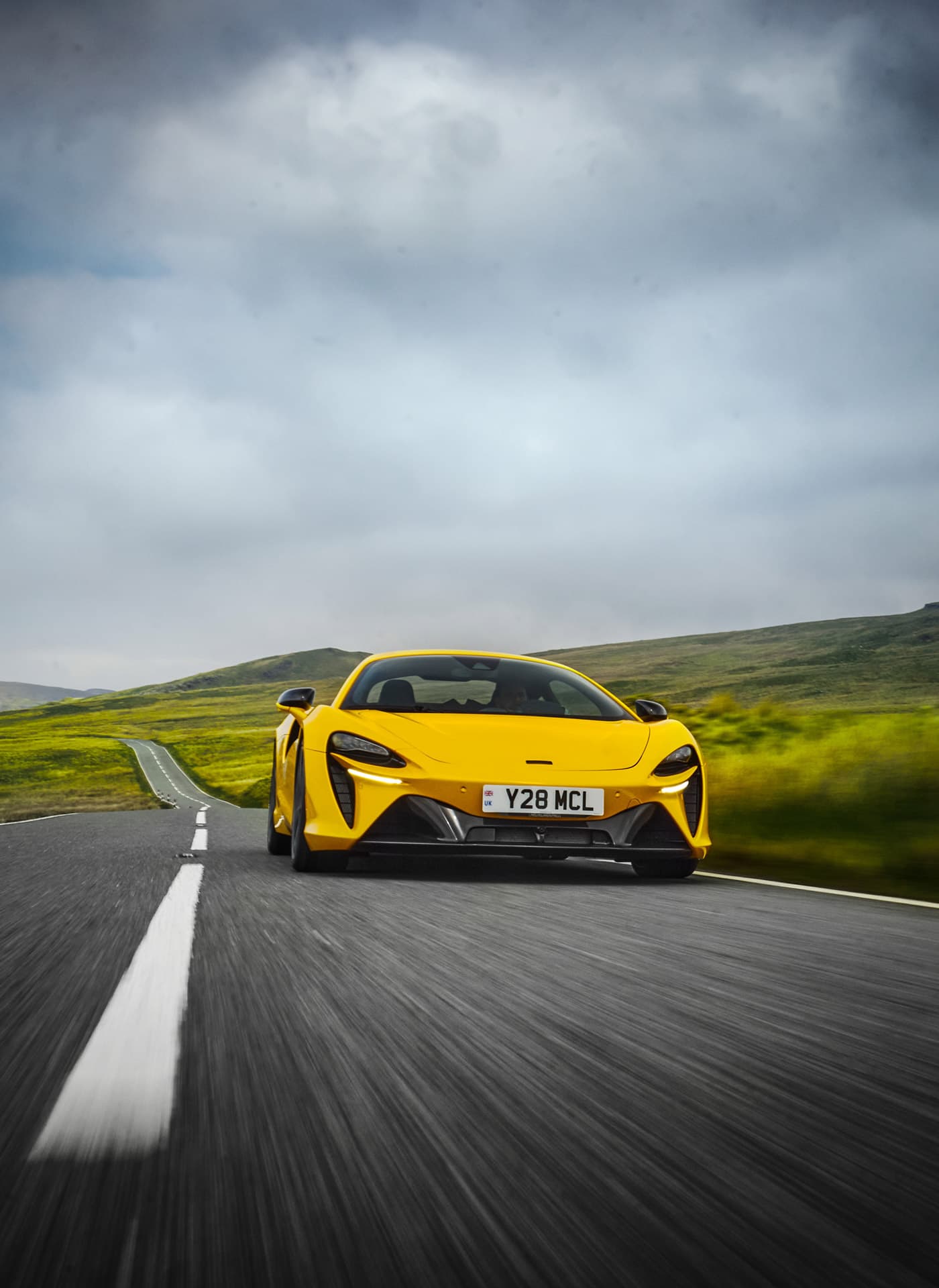 A dynamic front-on shot of a 2025 yellow McLaren Artura driving on a scenic road that stretches into a green, mountainous landscape.