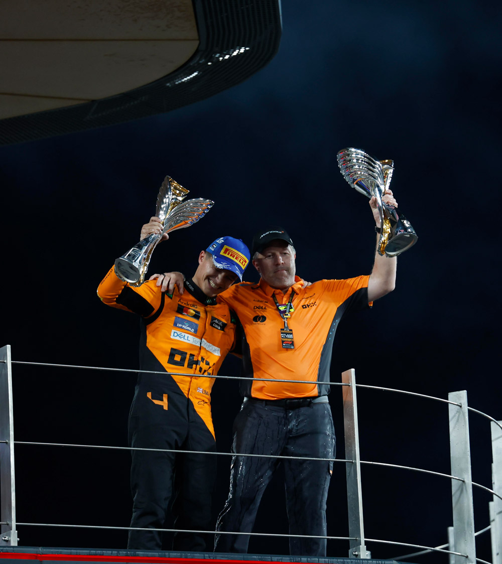 A jubilant photograph of McLaren F1 driver Lando Norris and CEO Zak Brown celebrating on a podium, each holding a trophy high in the air at a night race.