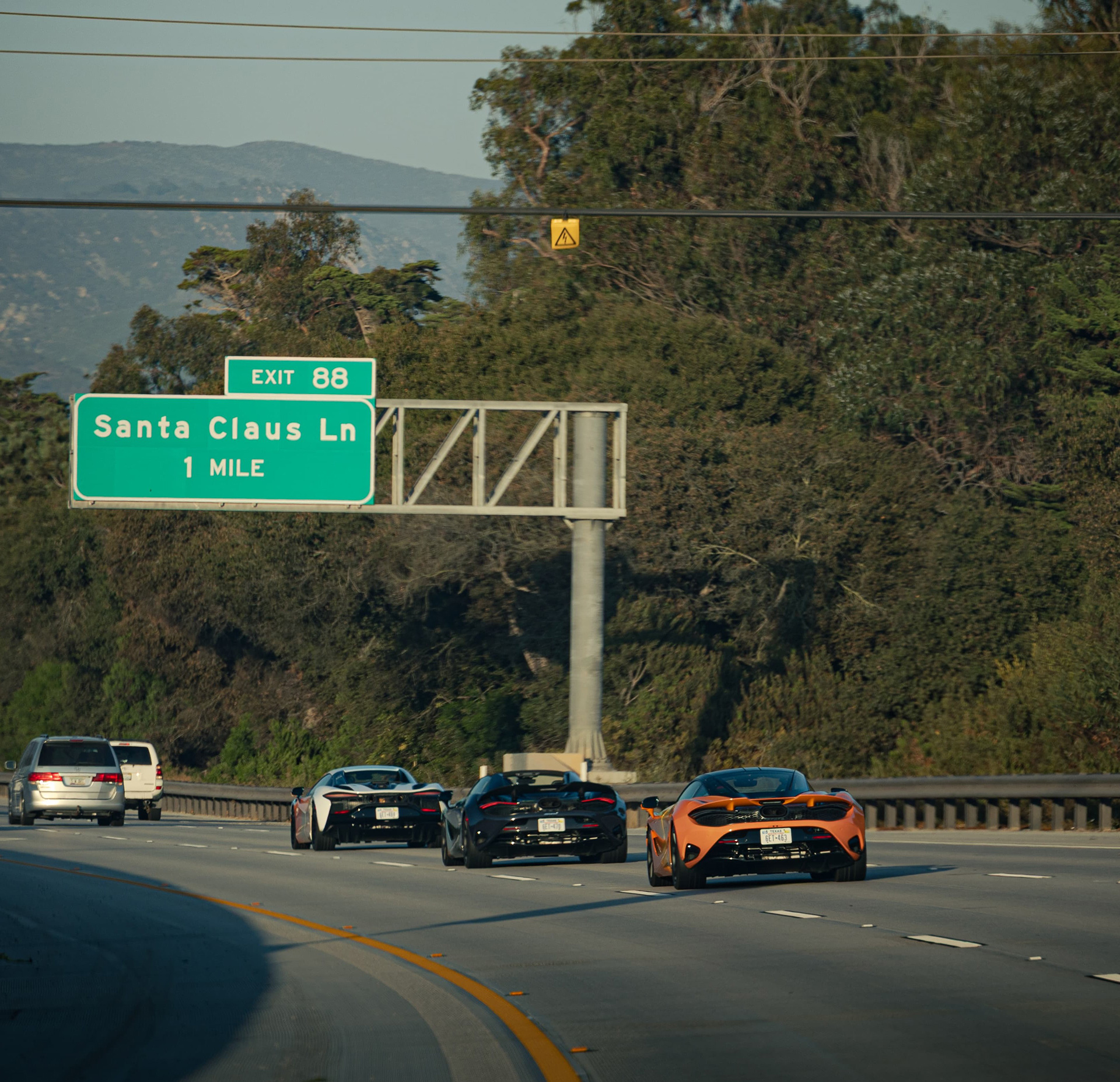 A convoy of McLaren supercars drives on a highway, with an overhead street sign for 'Santa Claus Ln' visible, as part of the 'States of Endurance' road trip.