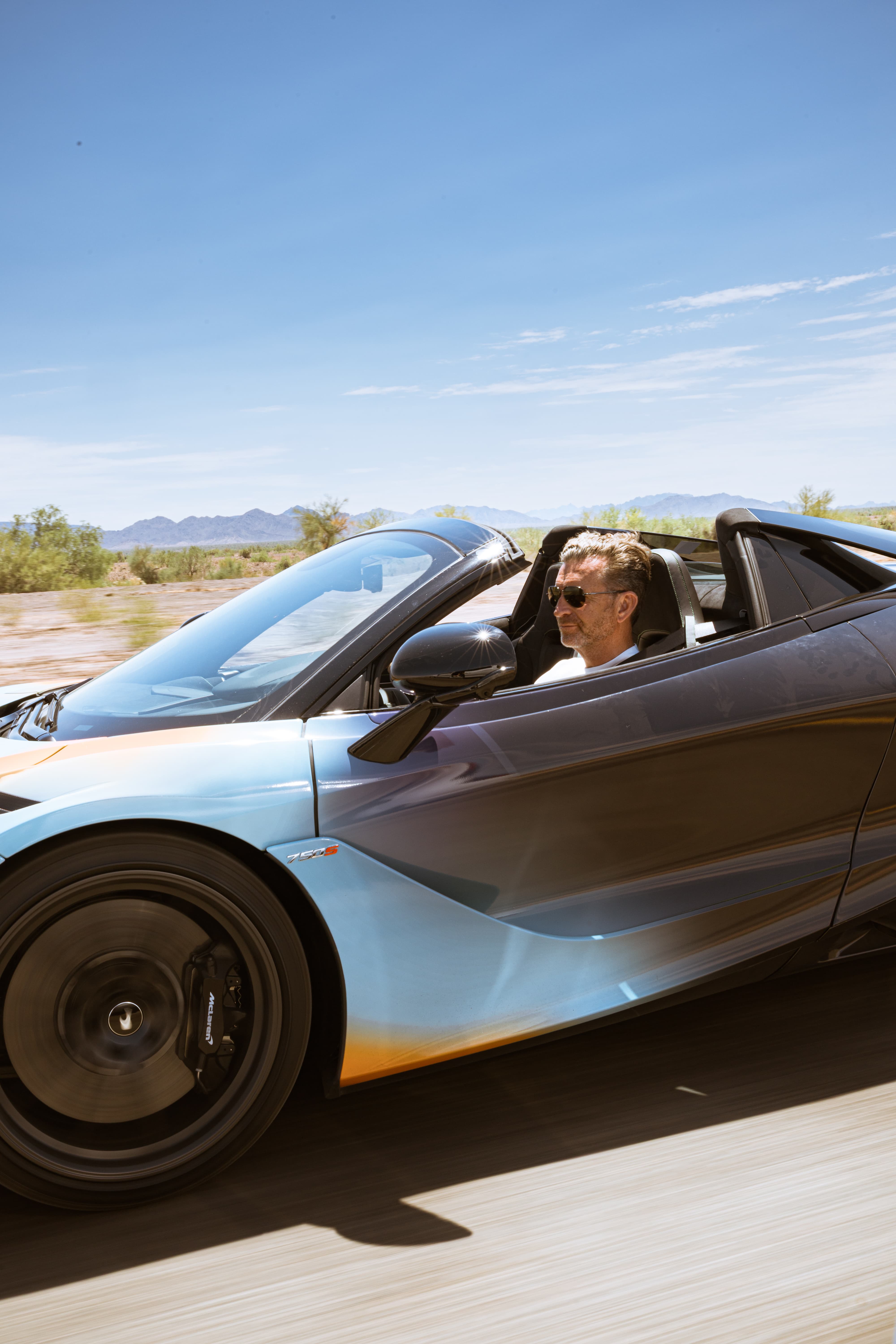 A man drives a McLaren convertible with a custom two-tone blue and orange livery through a sunny, arid landscape on the 'States of Endurance' road trip.