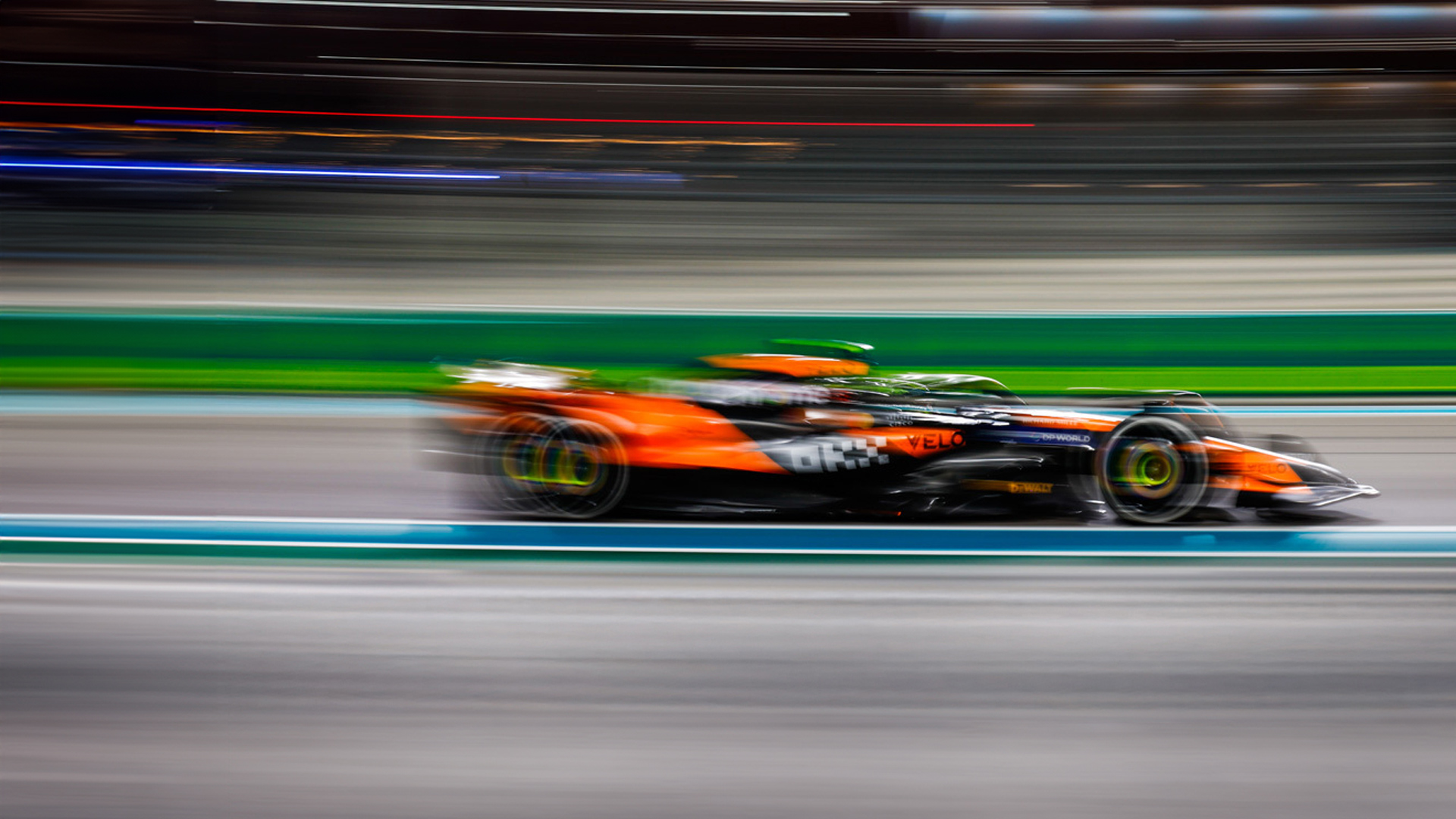 A dynamic, long-exposure shot of a McLaren Formula 1 race car in its orange and black livery speeding down a racetrack at night, with motion blur and streaks of light.