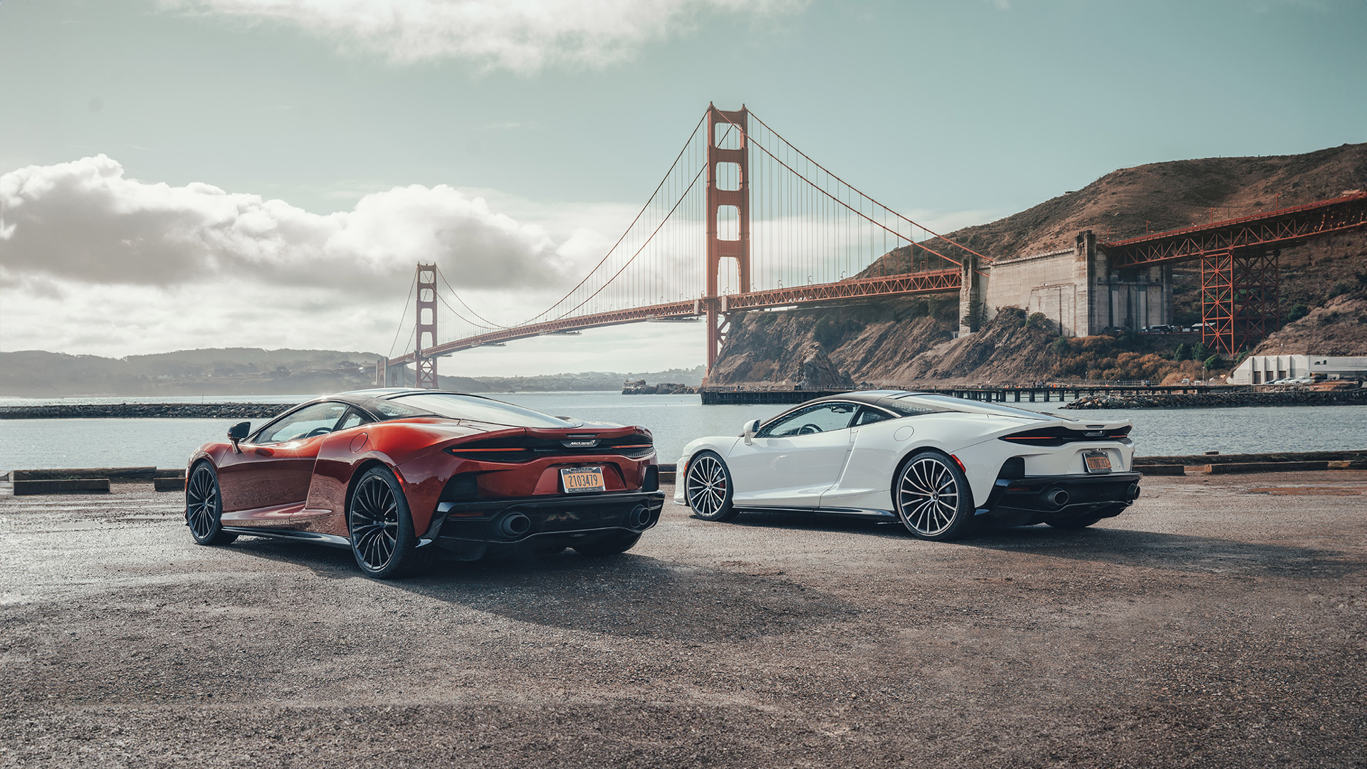A red and a white McLaren GT parked side by side with the iconic Golden Gate Bridge in San Francisco in the background.
