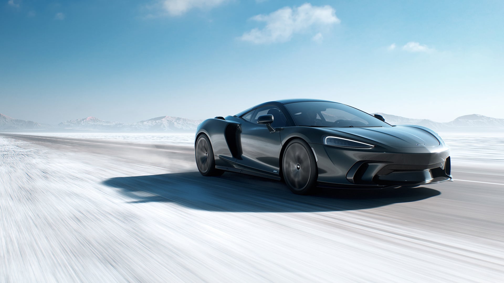 McLaren GTS high-performance sports car driving fast on a frozen, snow-dusted road. The sleek, dark grey GTS demonstrates its all-weather capability and precision-engineered power in the extreme winter landscape.