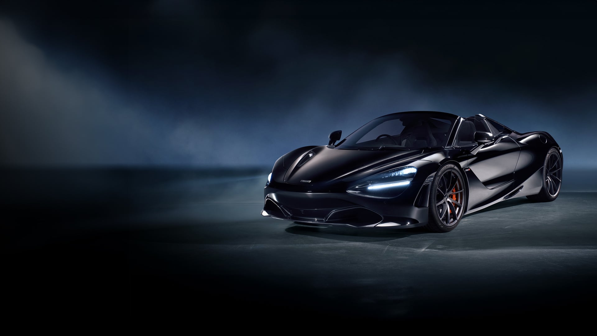A stunning black McLaren 720S Spider convertible is parked in a studio, showcasing its sleek front design, headlights, and orange brake calipers against a dark, moody background.