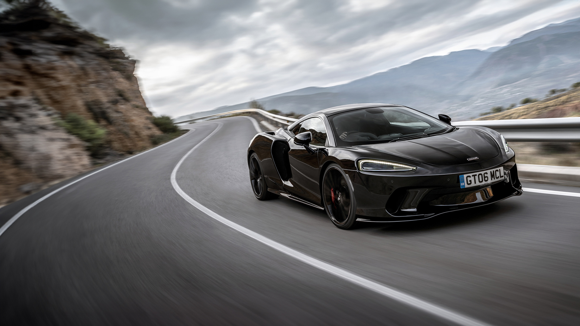  A black McLaren GT sports car drives on a winding road with mountains in the background, showcasing its sleek design and motion.
