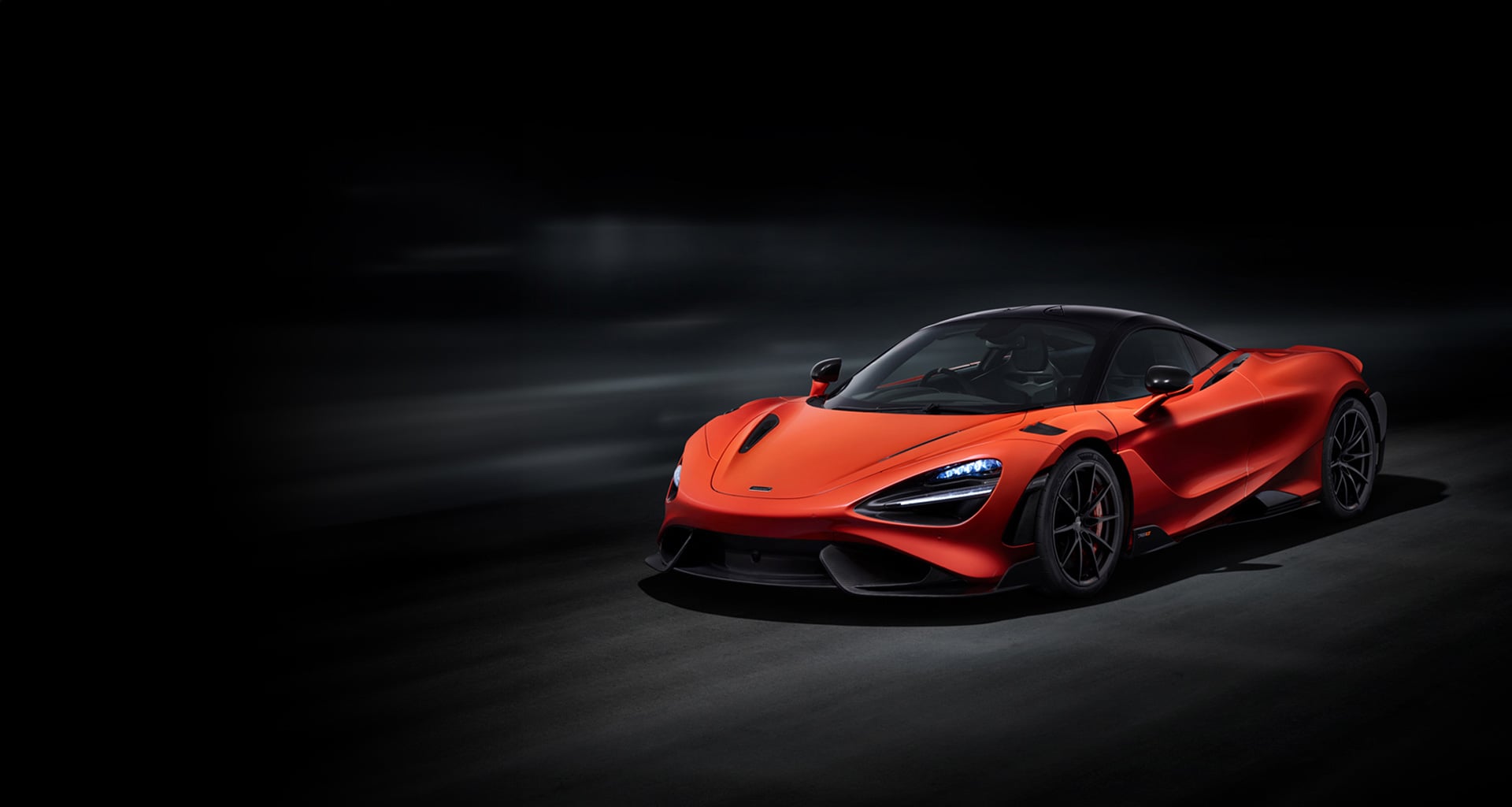 A vibrant orange McLaren 765LT supercar is shown from a low front-three-quarter angle in a dark studio setting. The car's sleek design and aggressive front are highlighted against the black background.