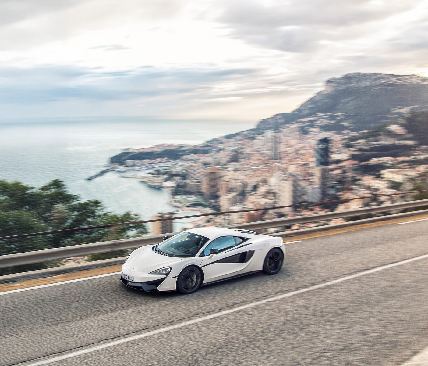 A white McLaren 540C driving along a cliff edge road at speed, during the day. Monte Carlo, Monaco can be seen blurry in the background.