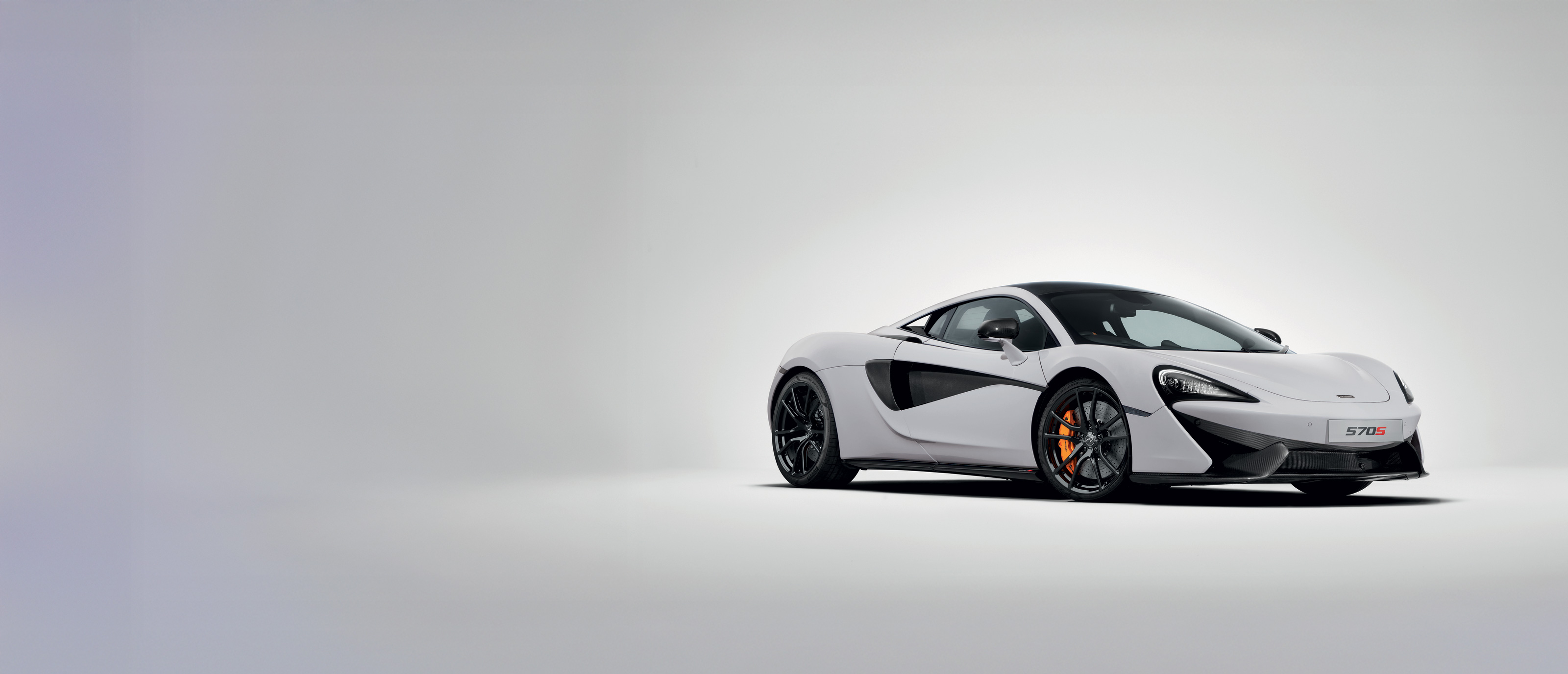 A white McLaren 570S sports car with orange brake calipers is shown from a front-three-quarter view on a white background.