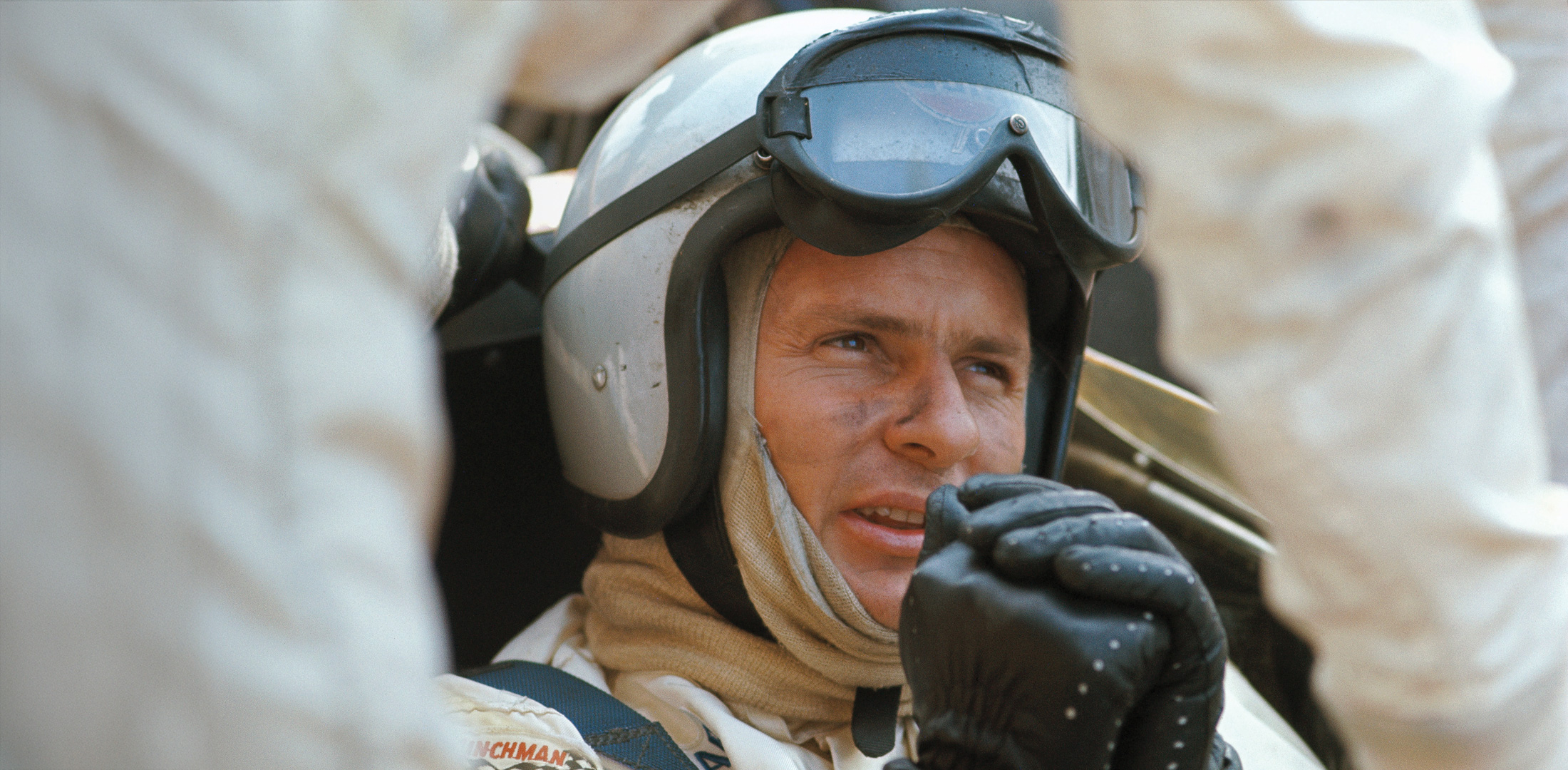 Close up of Bruce McLaren, sat in a racing car, wearing a helmet and driving gloves.