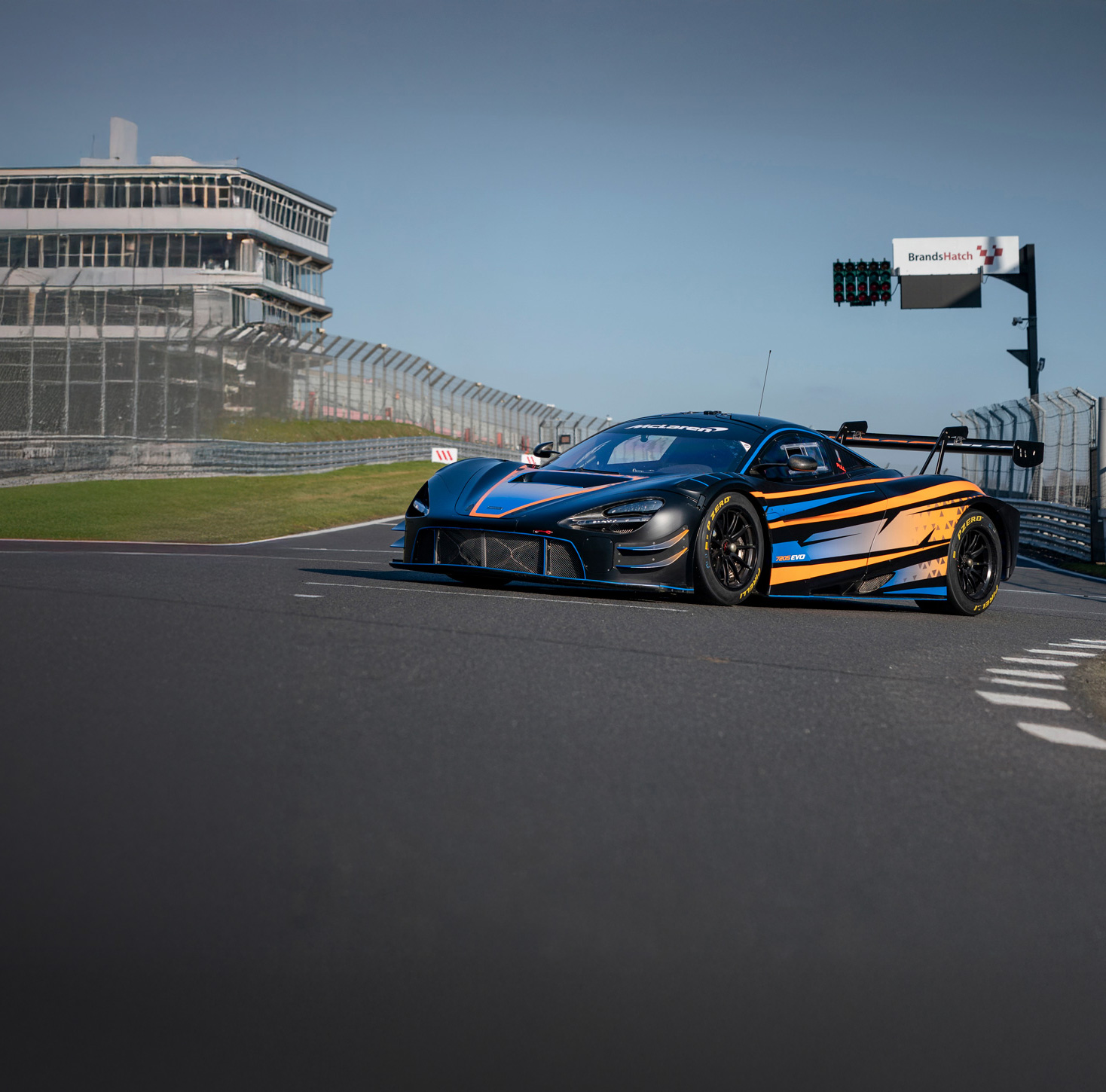 A black, orange and blue McLaren 720S GT3 Evo, parked across the start line of a race track, in the sun.