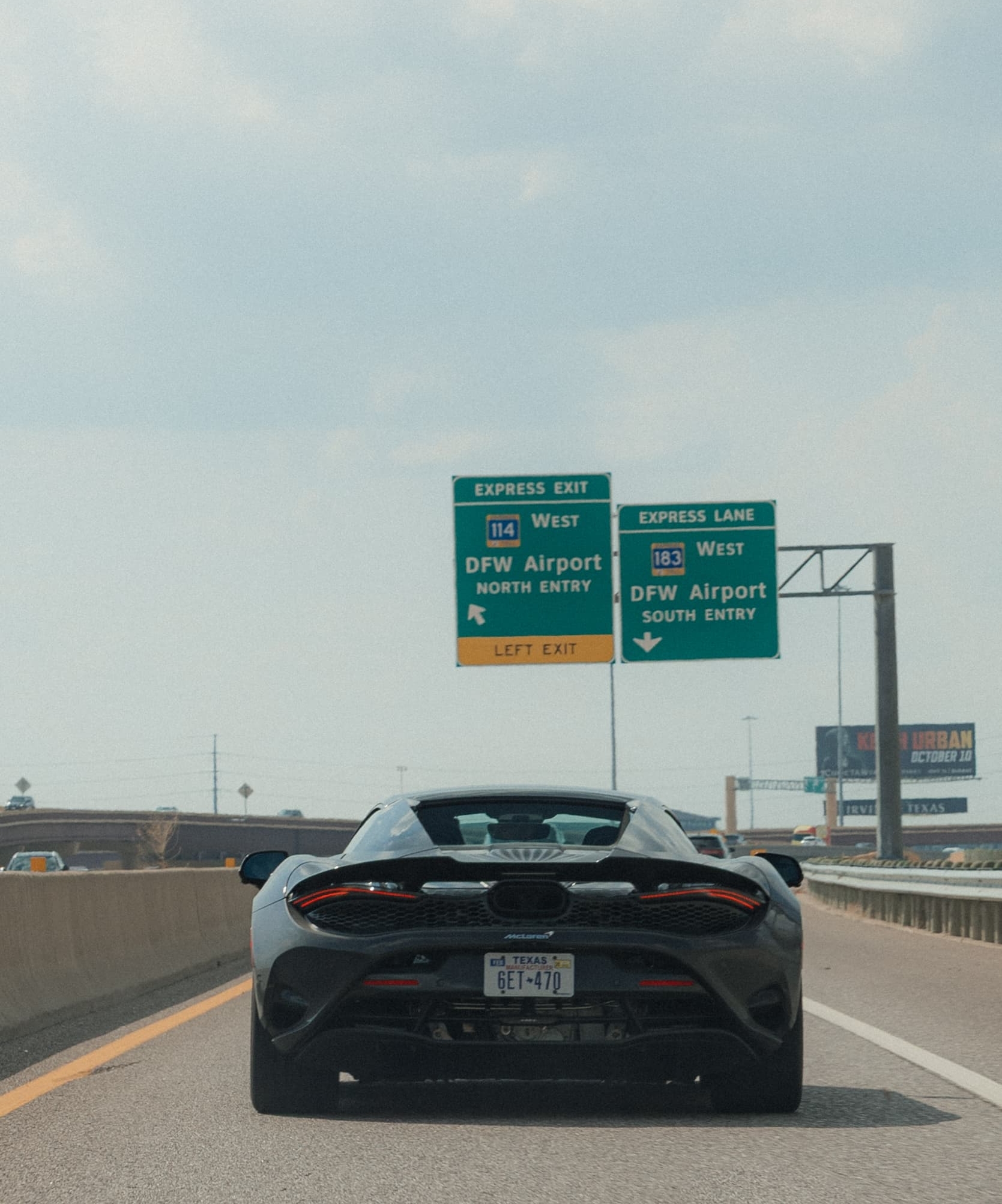 A McLaren driving under a green road sign which reads: 'express exit west, DFW Airport, north entry, left exit. A scene from Day 6 of the 'McLaren States of Endurance' road trip.