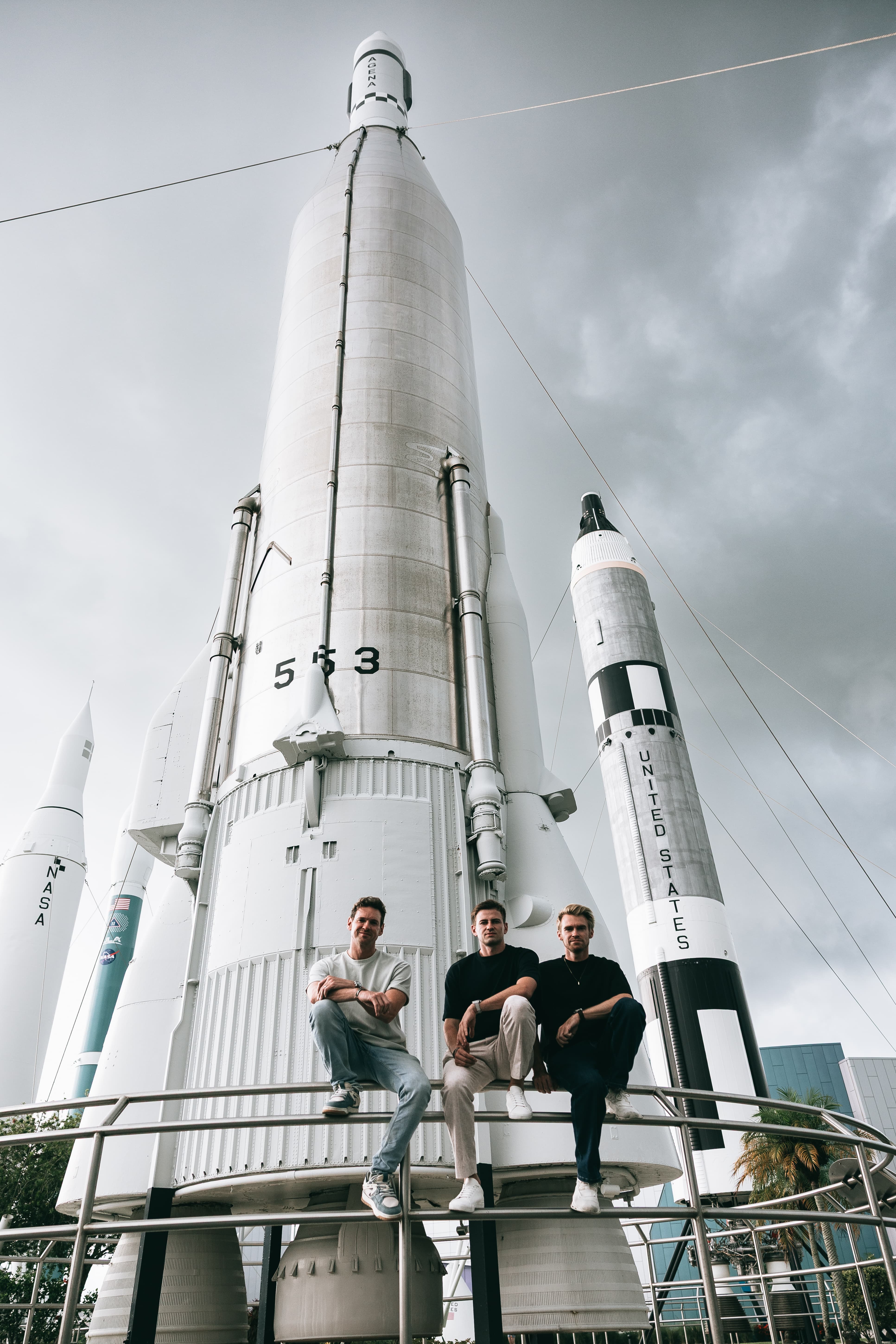 The McLaren States of Endurance team sat in front of a NASA rocket at the Kennedy Space Center, posing sat on the railing with their arms over their knees. A scene from Day 10 of the 'McLaren States of Endurance' road trip.