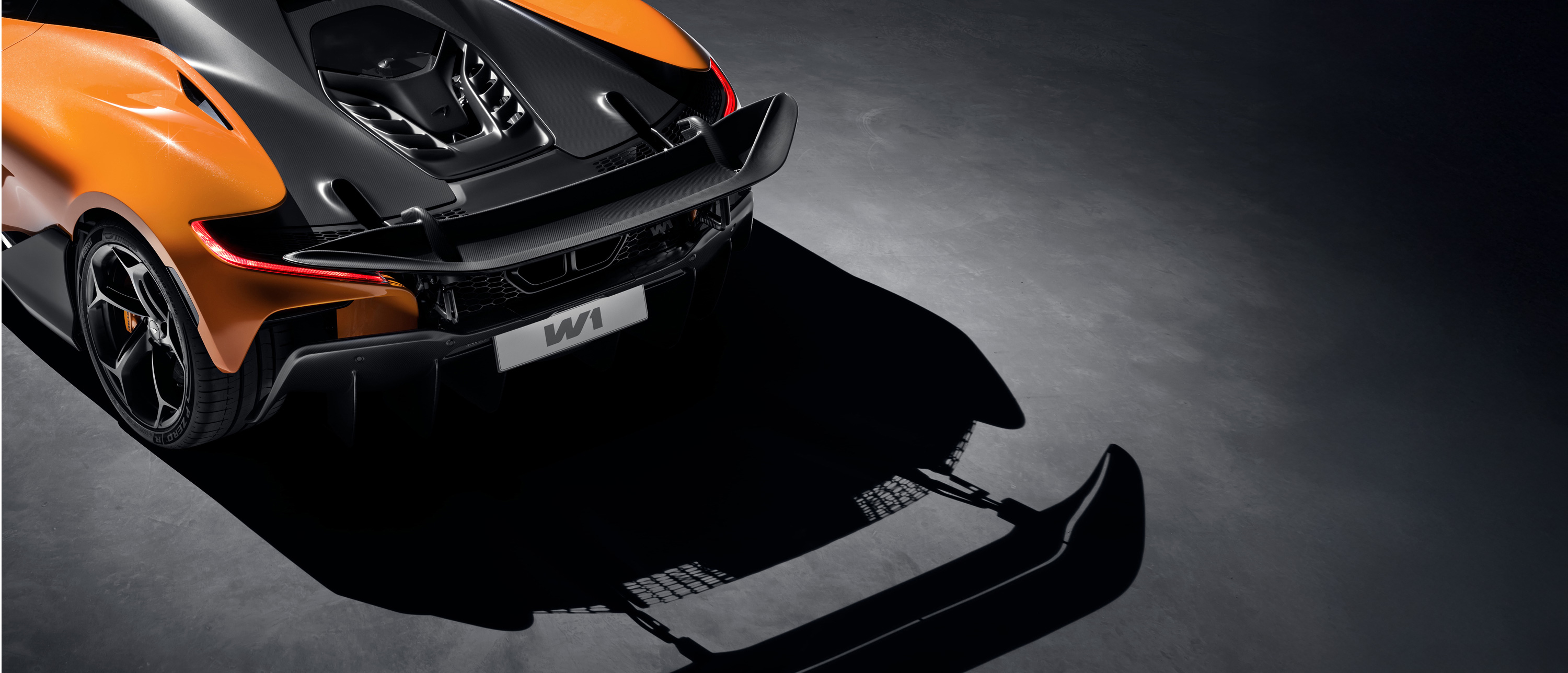A high-angle rear view of an orange and black McLaren supercar with its active rear wing deployed, casting a prominent shadow of the wing and diffuser on the dark ground below.