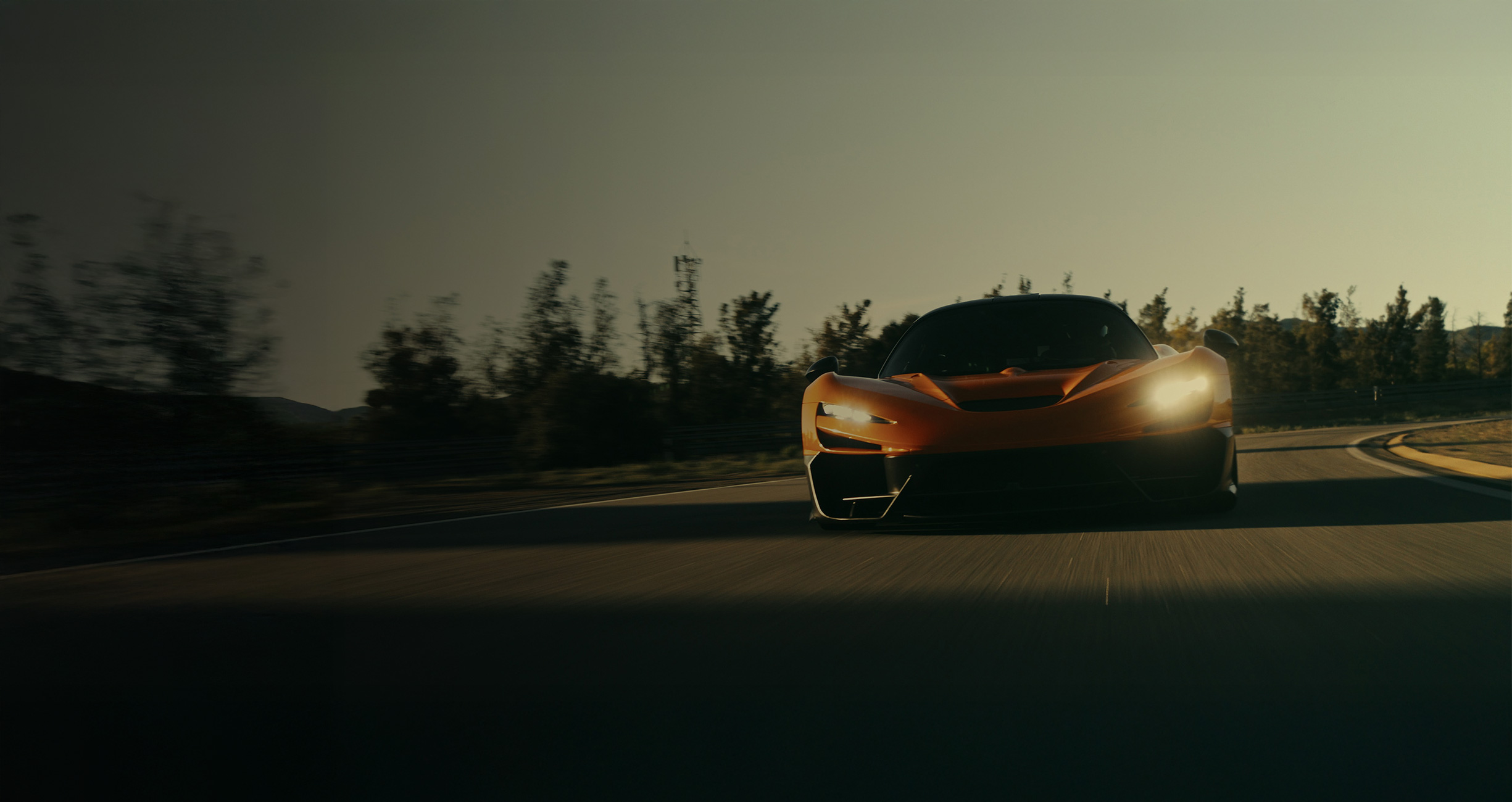  A low-angle, head-on shot of an orange McLaren supercar with its headlights on, speeding down a road under a dramatic, hazy sky with trees blurring in the background, conveying motion.