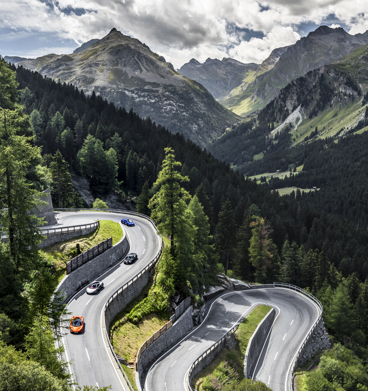 Series of McLaren cars driving down a winding mountain road surrounded by trees.