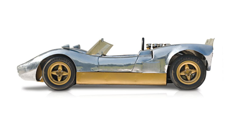 Side view of a silver/metal McLaren M1B, photographed on a red background.