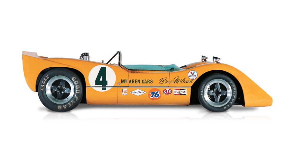 Side view of the orange McLaren M6A, with the number 4 painted on the side of the body and covered in sponsorship stickers, on a white background.