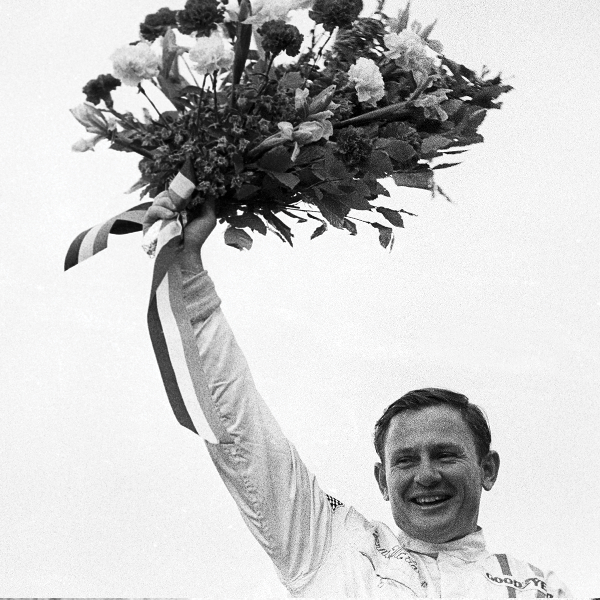 Black and white image of Bruce McLaren smiling and holding a bouquet of flowers in the air.