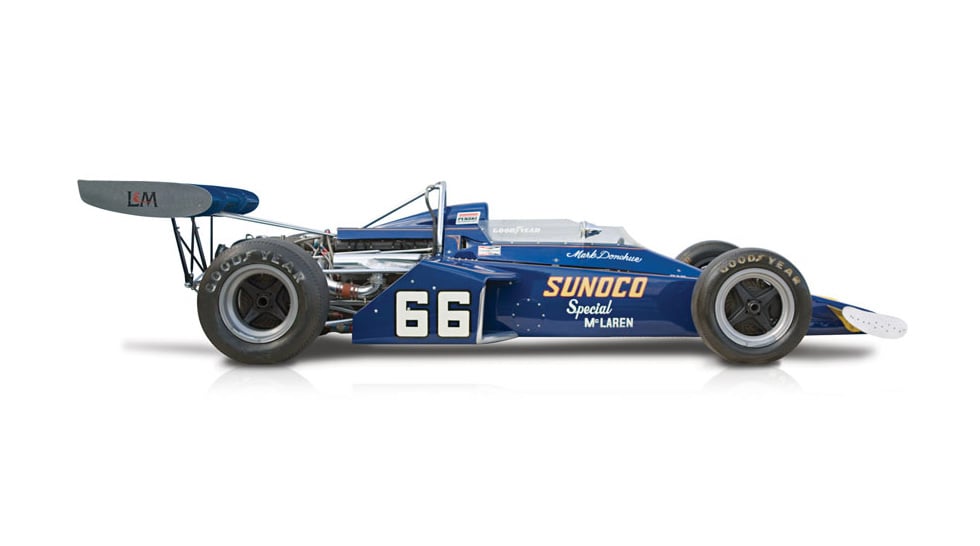 Side view of the blue McLaren M16 with the number 66 painted of the side of the body, and covered in sponsorship sticker. Shown on a white background.