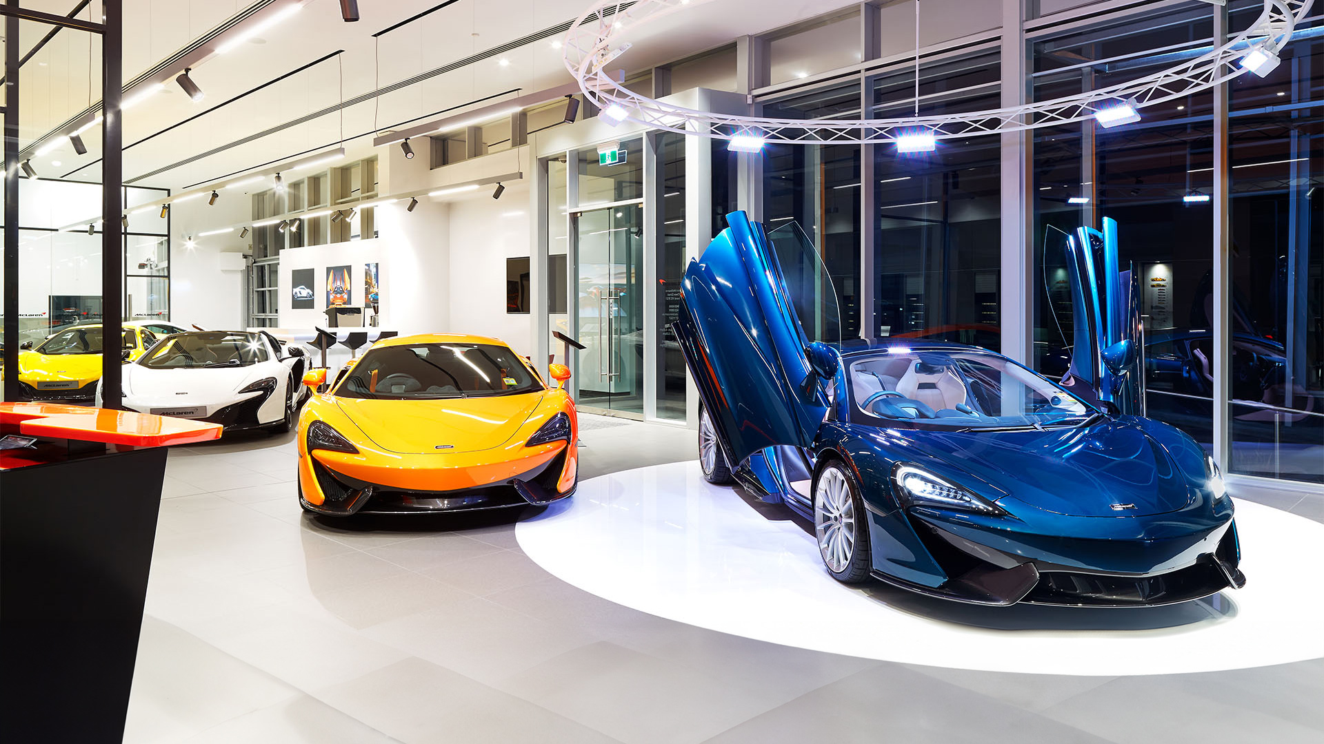 McLaren Melbourne showroom interior featuring a collection of supercars. A blue McLaren 570S/Artura convertible with its signature dihedral doors open is displayed next to an orange McLaren 570S/600LT coupe. Additional yellow and white McLaren sports cars are visible in the background.