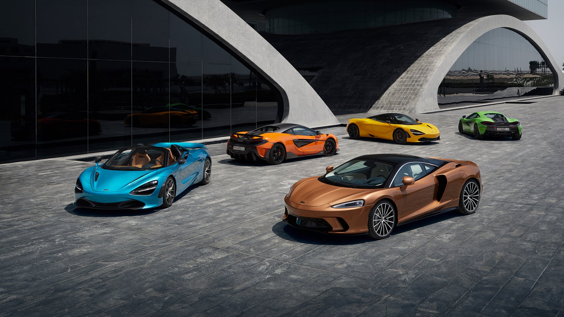 McLaren supercar model lineup exterior display featuring four cars in vibrant colours: Aurora Blue Artura Spider (convertible), McLaren Orange 750S, Volcano Yellow coupe, and a Bronze McLaren GTS. Showcases the full range of McLaren supercars and available exterior colours against modern architecture.