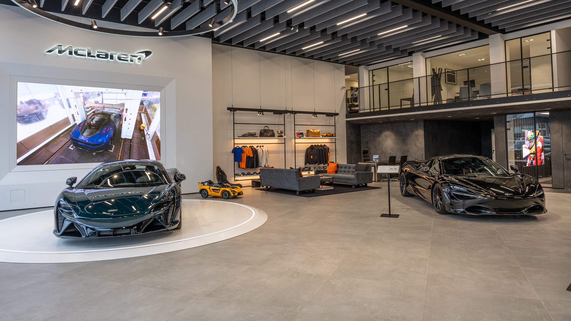 McLaren Brussels official retailer showroom featuring two luxury supercars: a dark green McLaren Artura Hybrid (left) on a rotating display, and a black McLaren 720S (right). The modern interior shows a comfortable customer lounge, branded apparel, and a large video display.
