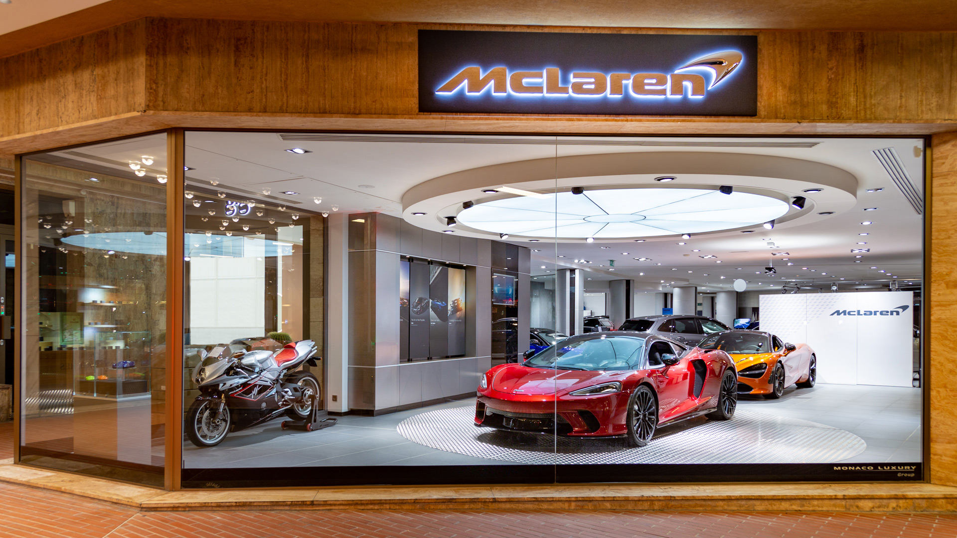 McLaren Monaco official retailer window display featuring a red McLaren 750S supercar and an orange McLaren in the background, alongside a high-performance exposed-engine motorcycle.