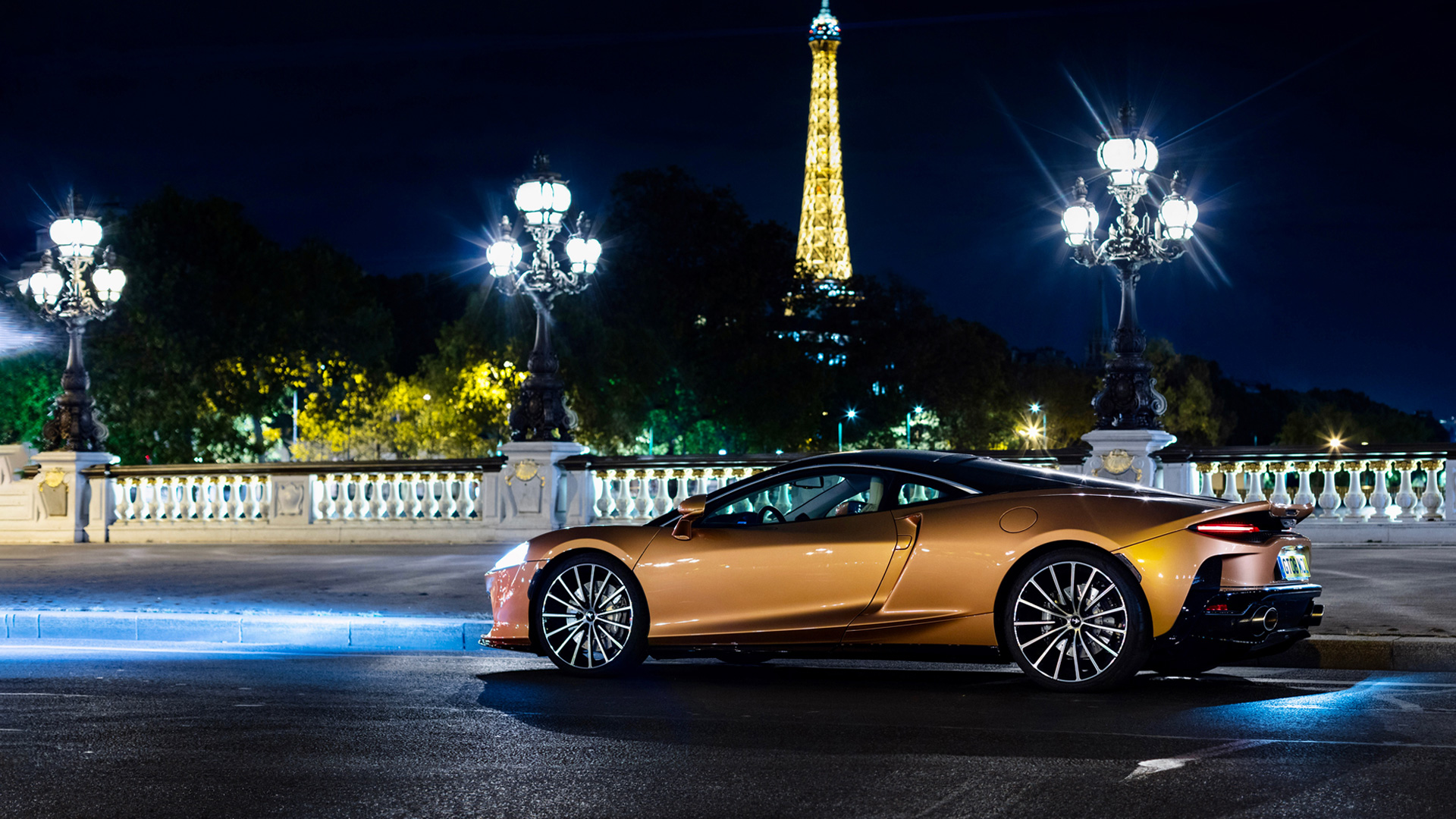 McLaren GT Grand Tourer (or GTS) in a unique Burnished Copper or bronze metallic paint, parked on a Parisian bridge at night. The illuminated Eiffel Tower and ornate street lamps are visible in the background.