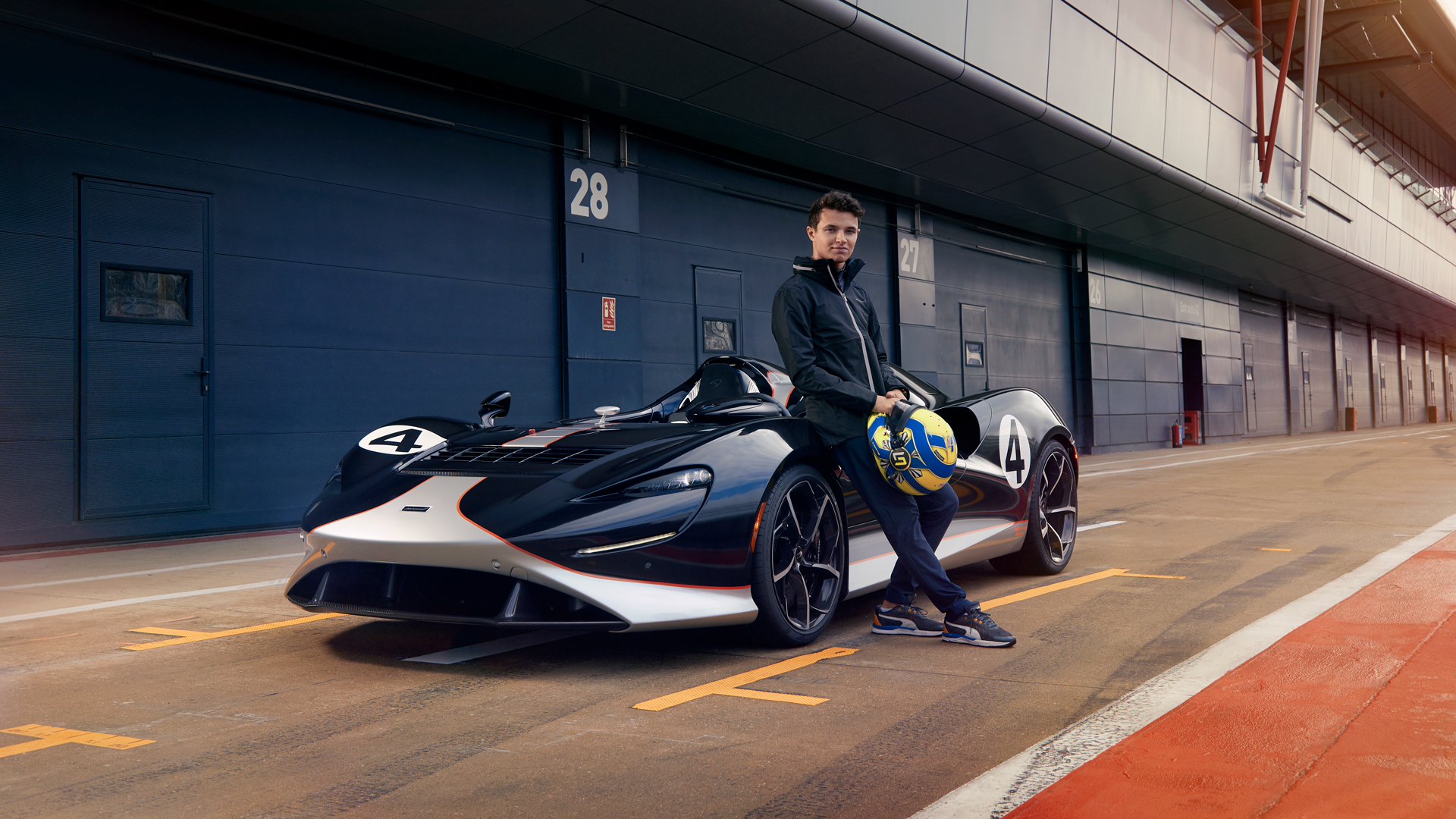 Lando Norris, McLaren F1 driver, posing with the exclusive McLaren Elva roadster in the pit lane at a circuit like Silverstone. The open-cockpit hypercar is finished in a custom black and silver livery with his race number 4, emphasizing Ultimate Series performance and F1 driver collaboration.