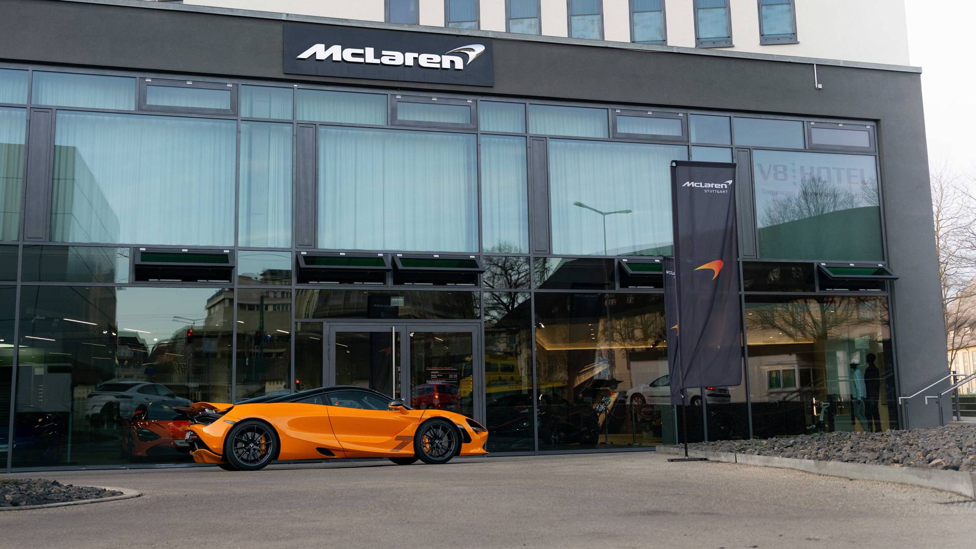 McLaren Stuttgart official retailer exterior at Motorworld Böblingen, Germany. A bright McLaren Orange 750S supercar is parked outside the modern glass showroom, with the McLaren signage visible above.