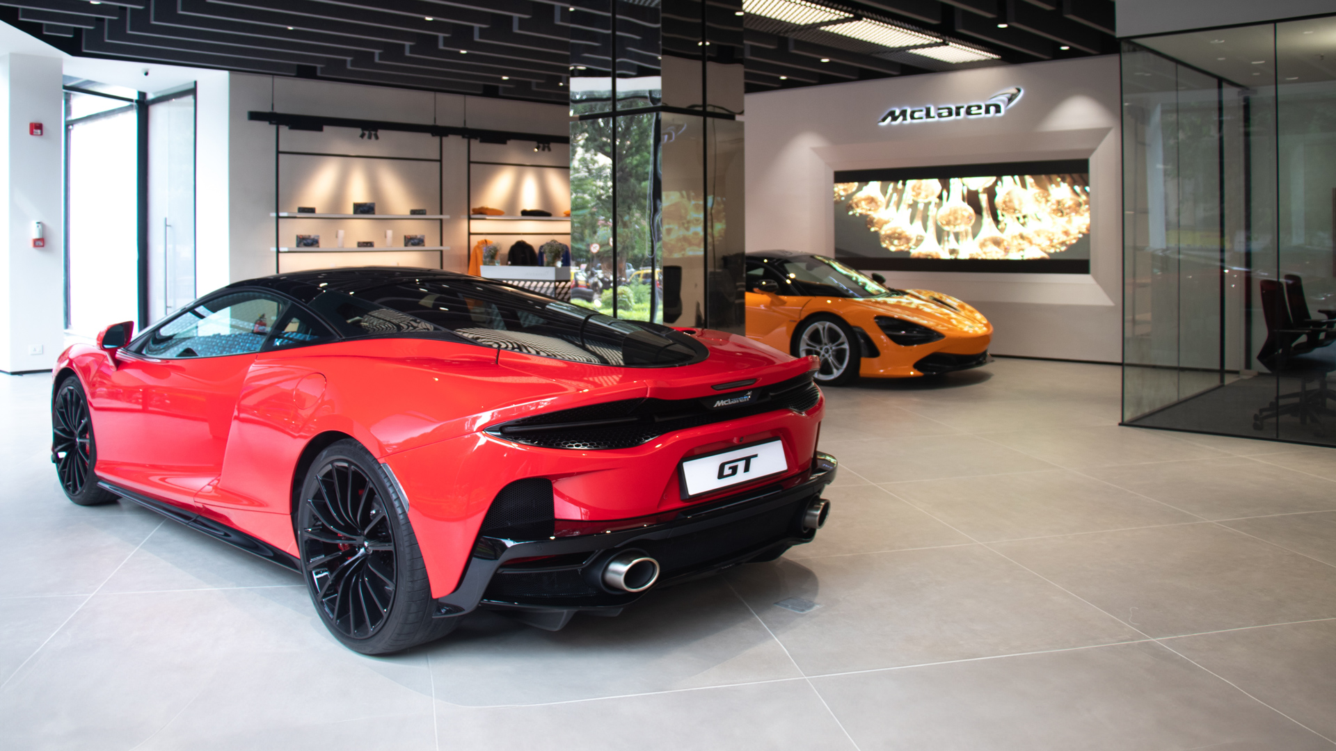 McLaren Mumbai official retailer showroom interior. A red McLaren GT Grand Tourer is parked in the foreground, with an orange McLaren 750S supercar in the background.