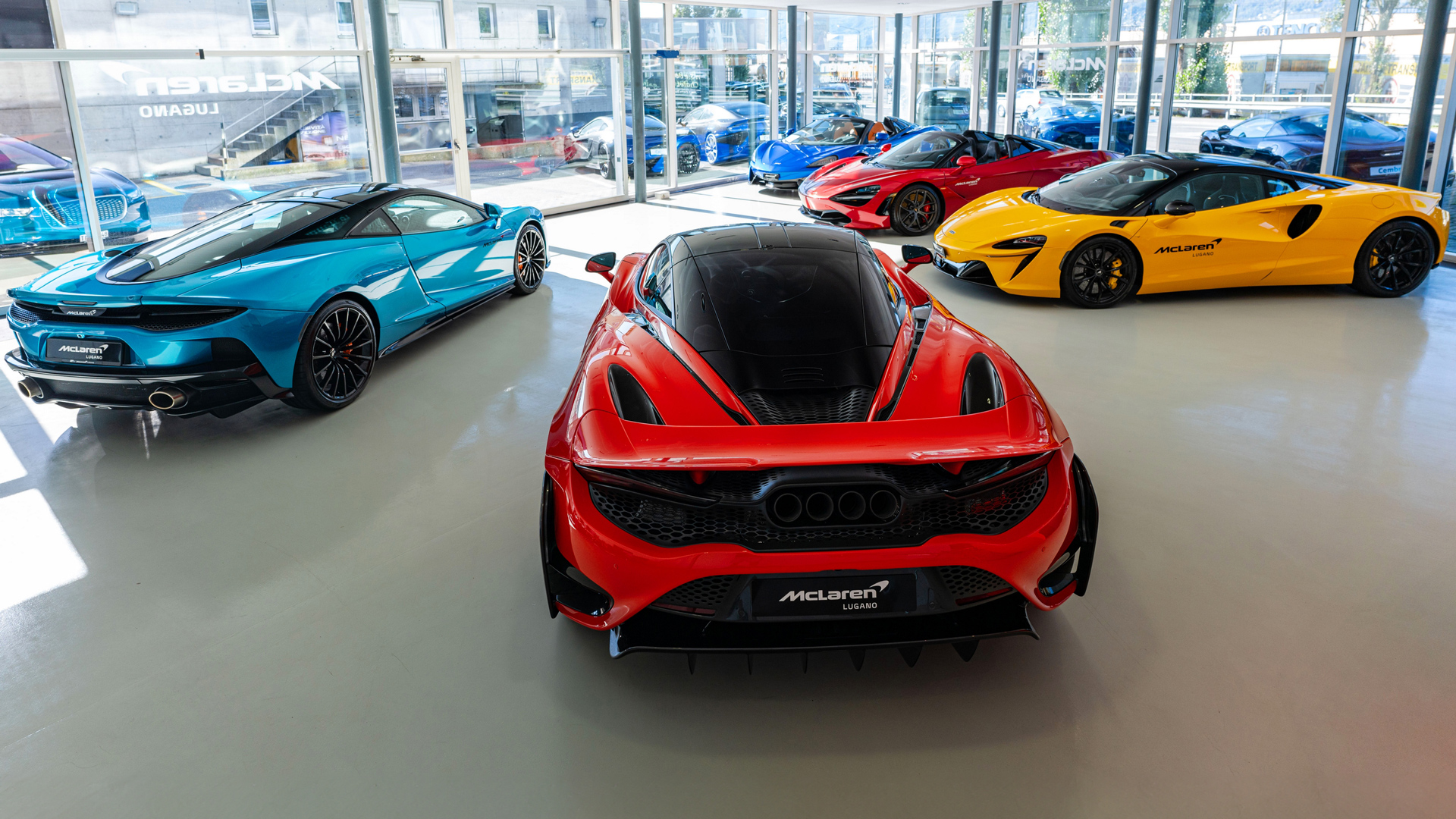 McLaren Lugano, Switzerland retailer showroom showcasing multiple luxury supercars. The display features a Red McLaren 750S (center rear), a Blue McLaren GT, and a Yellow McLaren Artura model.