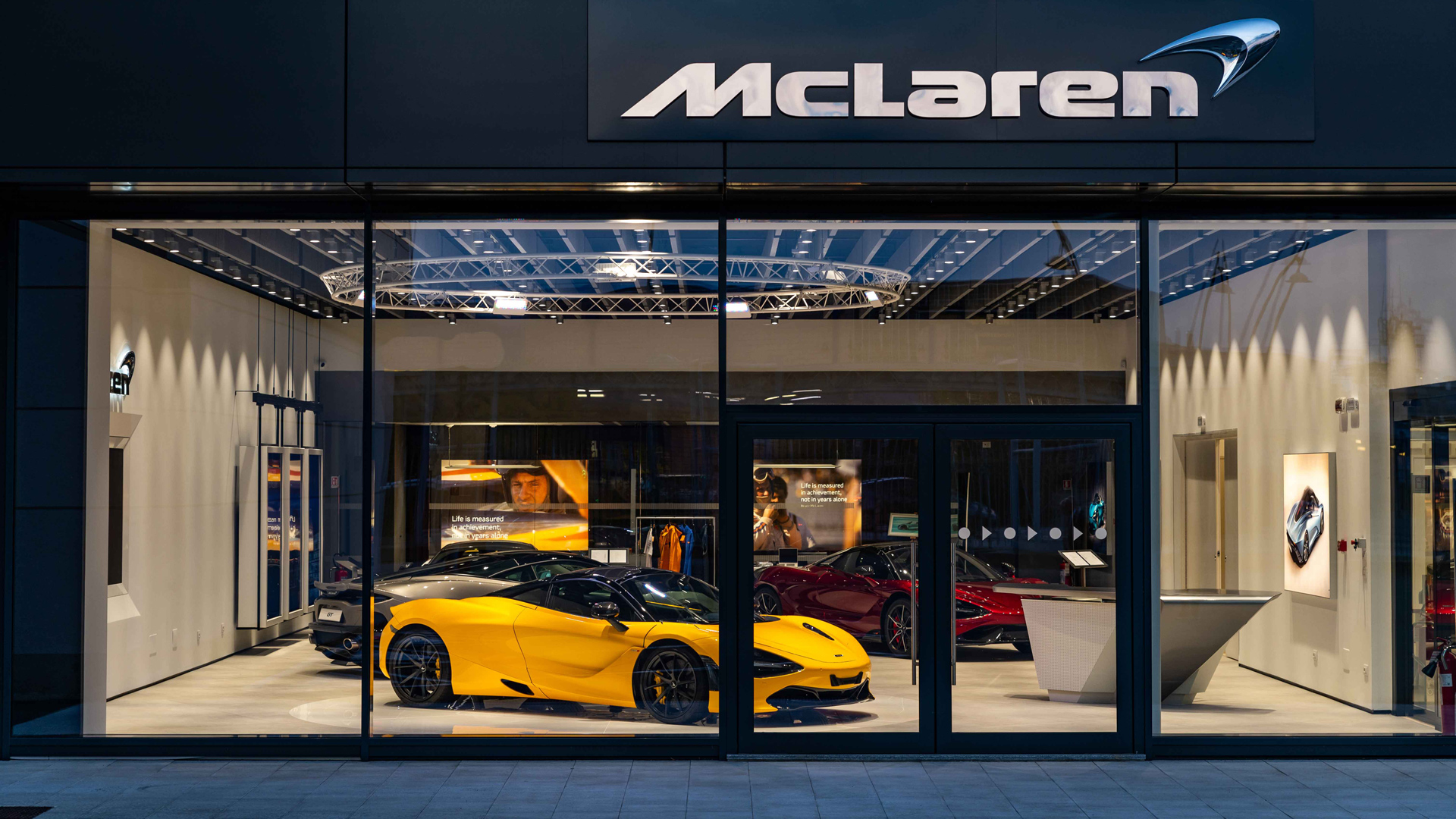 McLaren official retailer showroom exterior at night, featuring a vibrant Yellow McLaren 720S supercar displayed prominently in the front window.