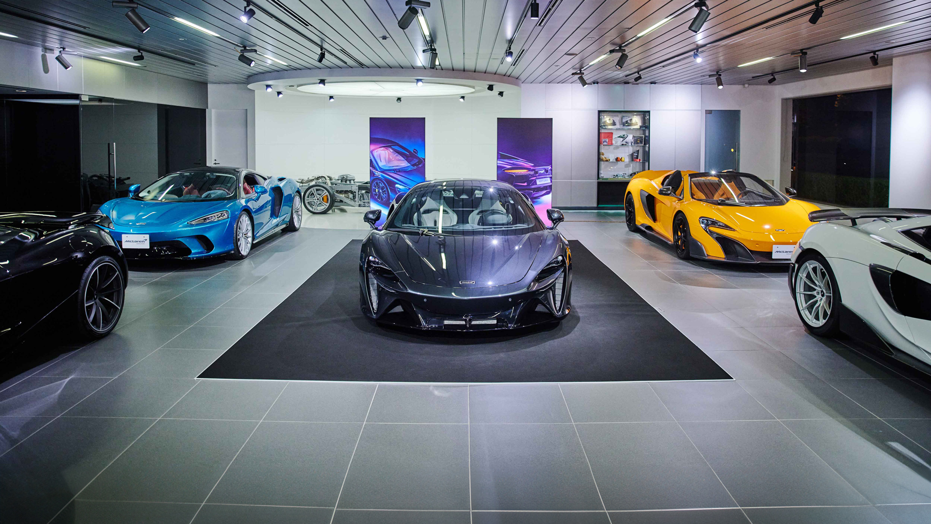 McLaren Tokyo official retailer showroom featuring a multi-car display. A dark grey McLaren Artura is spotlighted, flanked by a blue 750S and a yellow P1 supercar.
