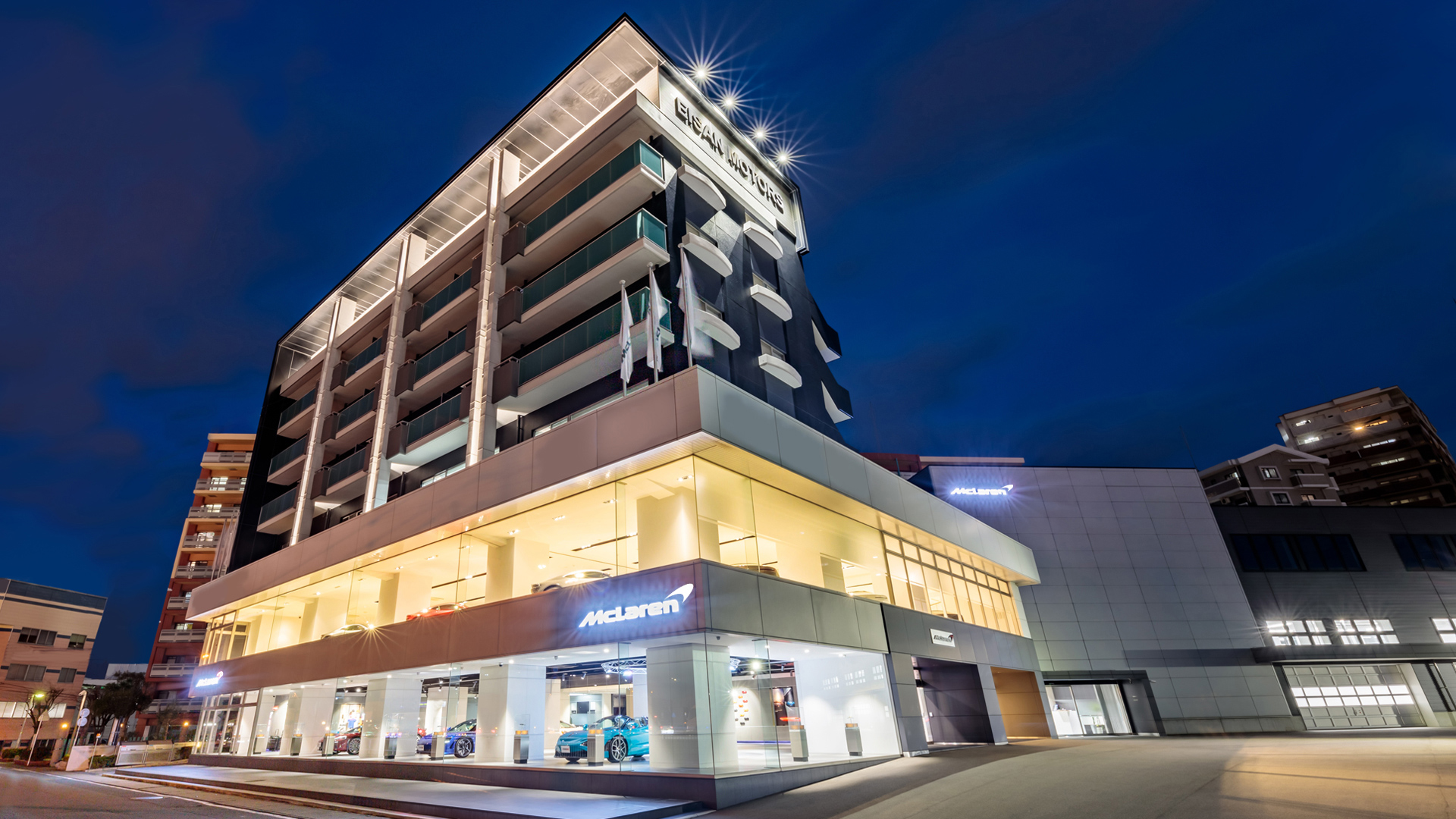 McLaren Fukuoka official retailer exterior at night in Japan. The modern, multi-story glass and concrete building features the McLaren logo prominently, with luxury supercars visible on the brightly lit ground-floor showroom.