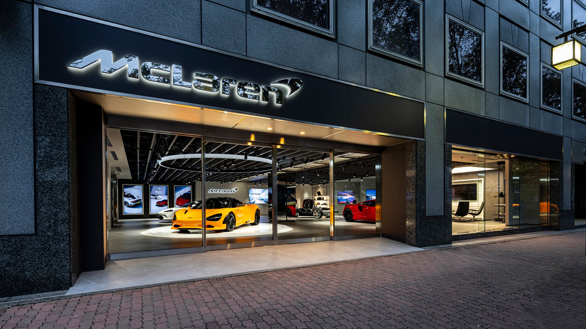 McLaren Nagoya official retailer exterior at night in Japan. The modern, illuminated showroom features a vibrant Yellow McLaren 720S in the display window, flanked by red and white supercars.