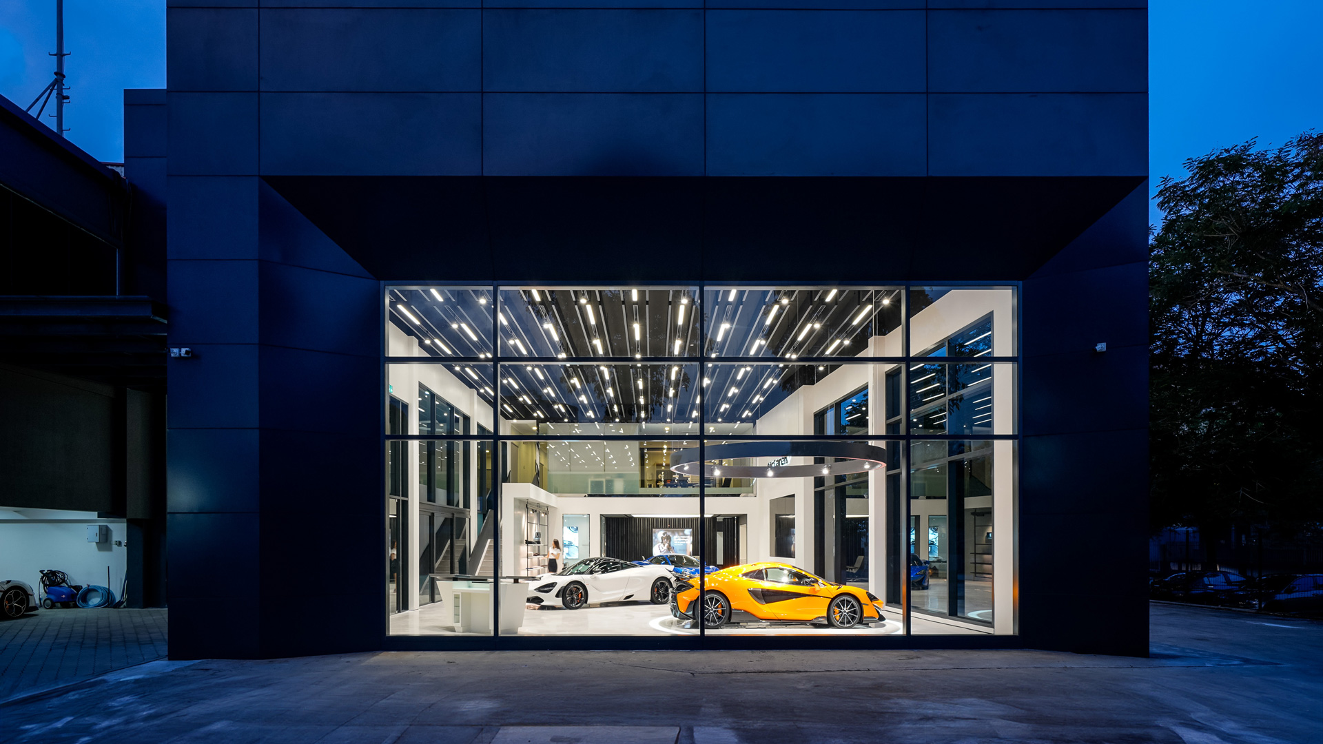 McLaren Kuala Lumpur official retailer exterior at dusk in Malaysia. The sleek, modern black building features a wide glass window showcasing a McLaren Orange supercar and a white McLaren model inside.