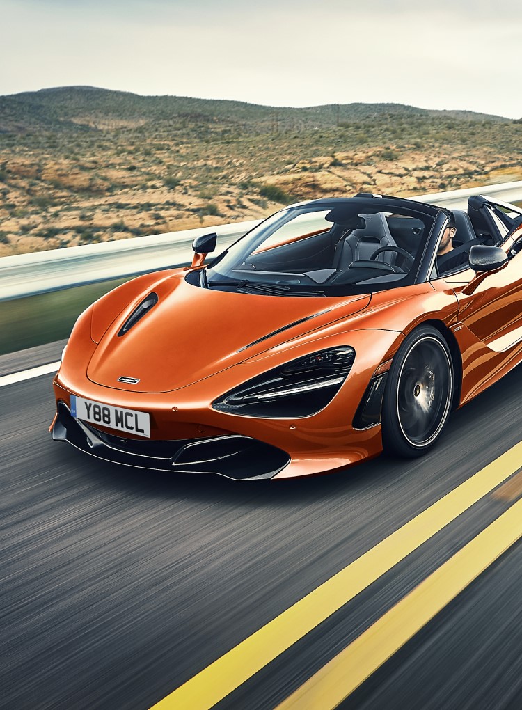 A front-side view of the McLaren 720S Spider convertible in a vibrant orange, driving on a road surrounded by a arid, rural landscape.