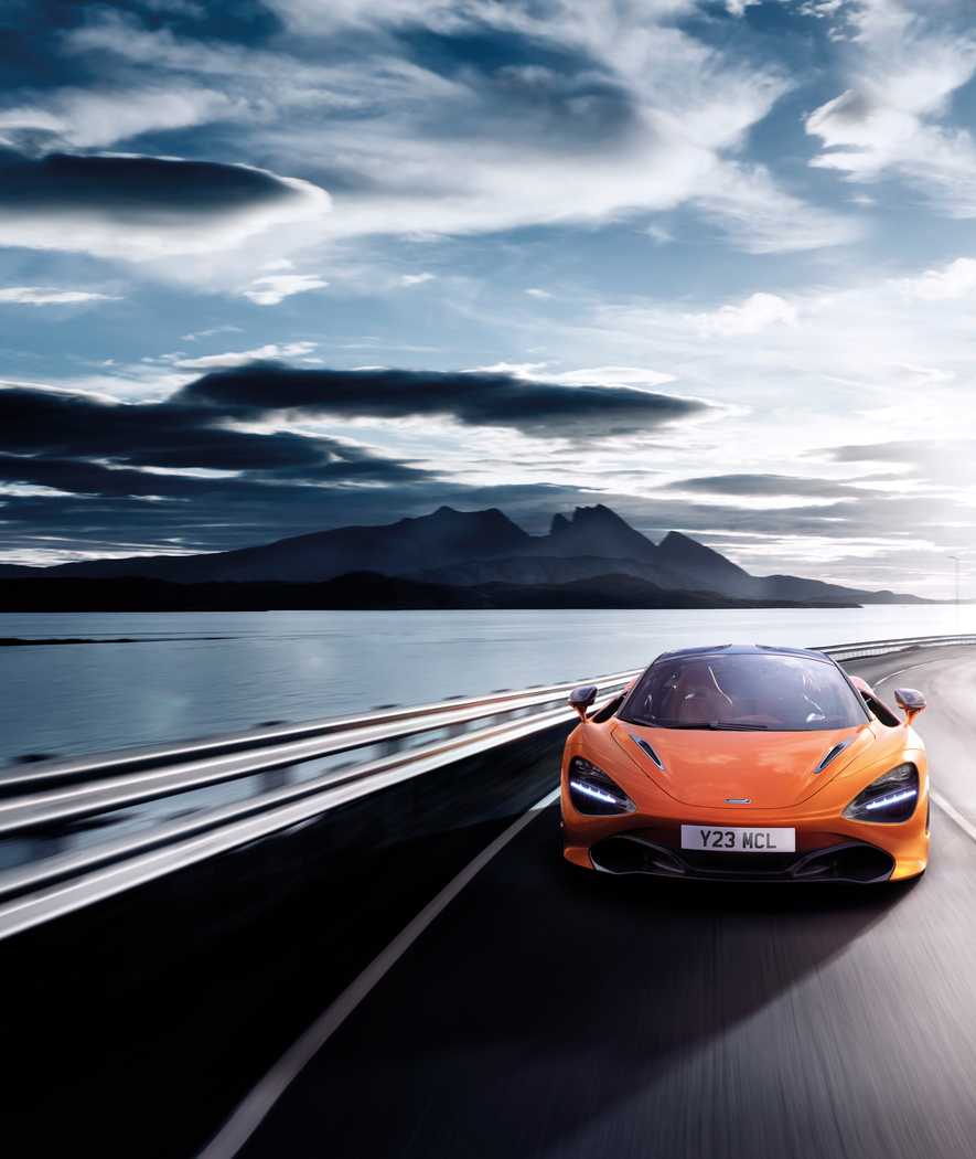 The McLaren 720S, a high-performance supercar, driving at speed on a winding coastal road with mountains and water in the background.