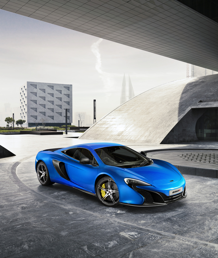 The McLaren 650S in a bright blue finish, positioned in a modern urban plaza with unique buildings in the background.