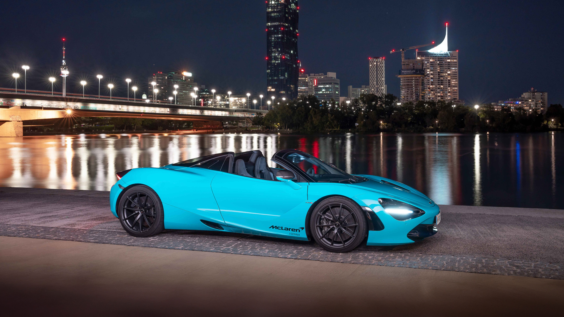 McLaren 720S Spider convertible in a vibrant Ceramic Blue or Belize Blue paint, parked against the illuminated Vienna skyline at night. Features the Danube Tower and modern architecture.