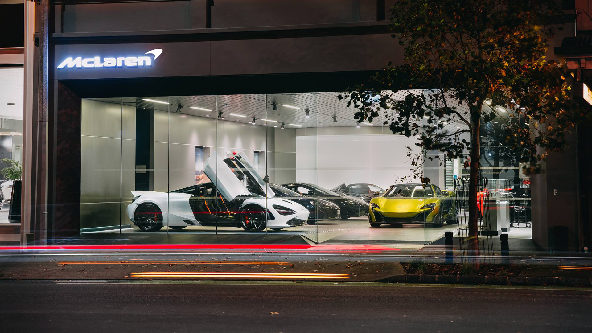 Night-time exterior view of the McLaren Auckland supercar showroom with multiple McLarens on display, including a white McLaren 720S with dihedral doors open, and street traffic light trails.