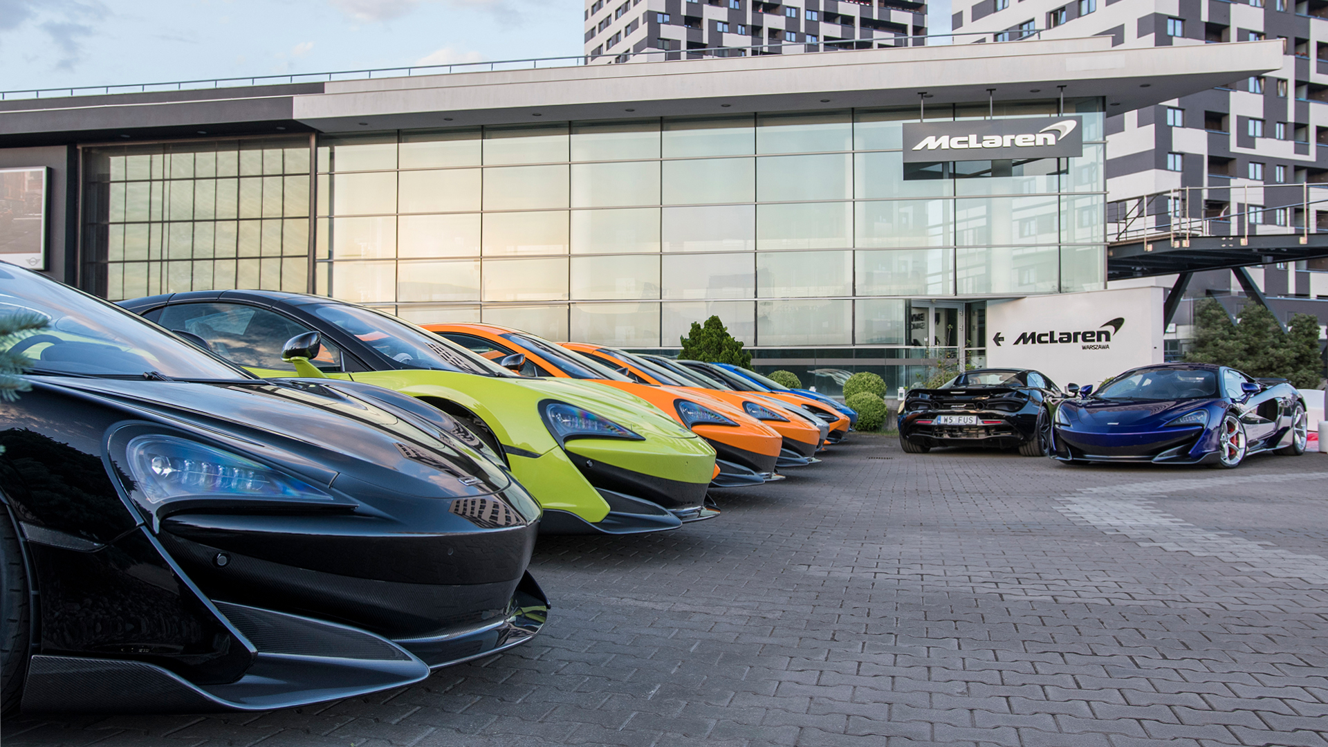 A row of colorful McLaren supercars, including yellow, orange, and blue models, parked outside the modern McLaren dealership and showroom, highlighting a vibrant model lineup.
