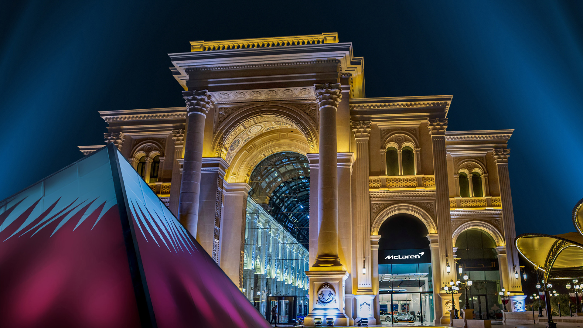 Exterior night view of the stunning McLaren Doha showroom in Qatar, set beneath a grand, illuminated classical archway and glass atrium, representing luxury supercar retail in the Middle East.